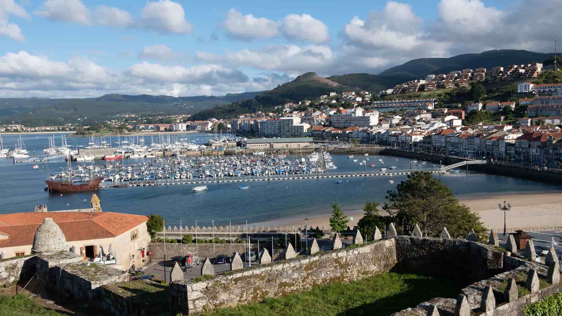 An elevated view of the picturesque harbor of Baiona, Spain, from behind a historic stone wall. The bay is filled with dozens of sailboats and yachts, while the town's white and red-roofed buildings climb the surrounding green hills.