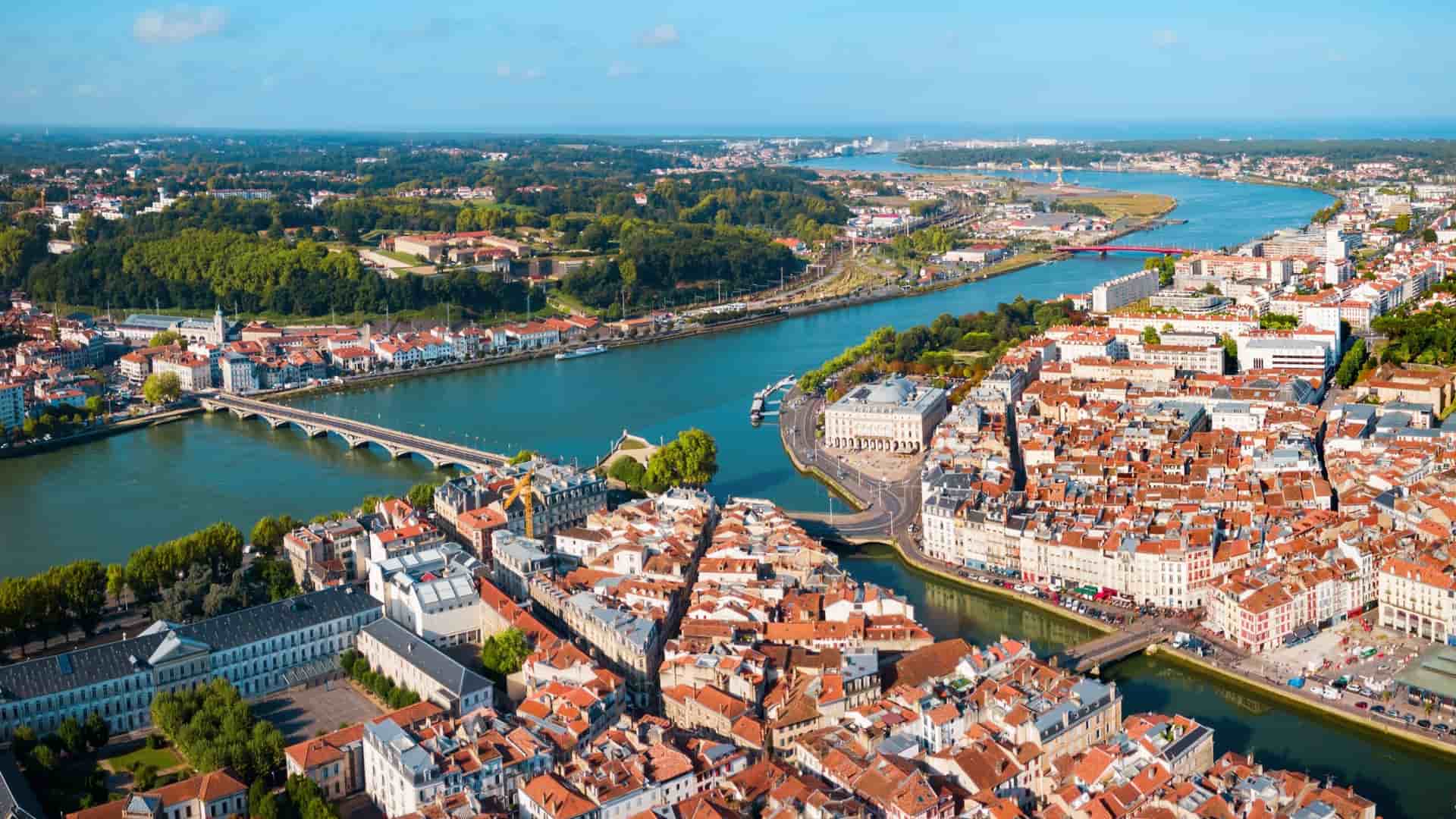 An aerial view of Bayonne, France, showing the colorful houses, river, and bridges under a clear blue sky, extending toward the ocean horizon.
