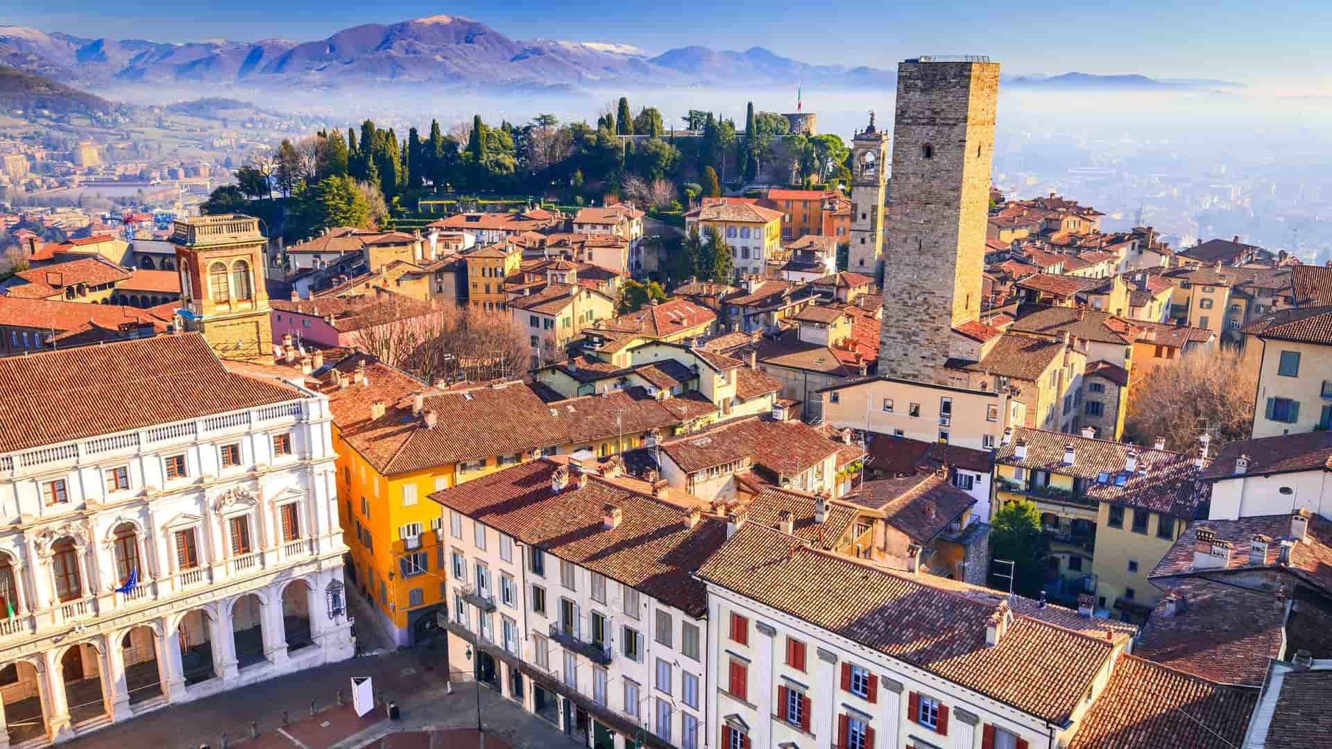 An elevated panoramic view of the historic Città Alta of Bergamo, Italy. The foreground is a dense collection of buildings with traditional red tile roofs, centered around a tall, medieval stone tower. The city spreads into the distance, with a range of snow-dusted mountains on the horizon under a clear blue sky.