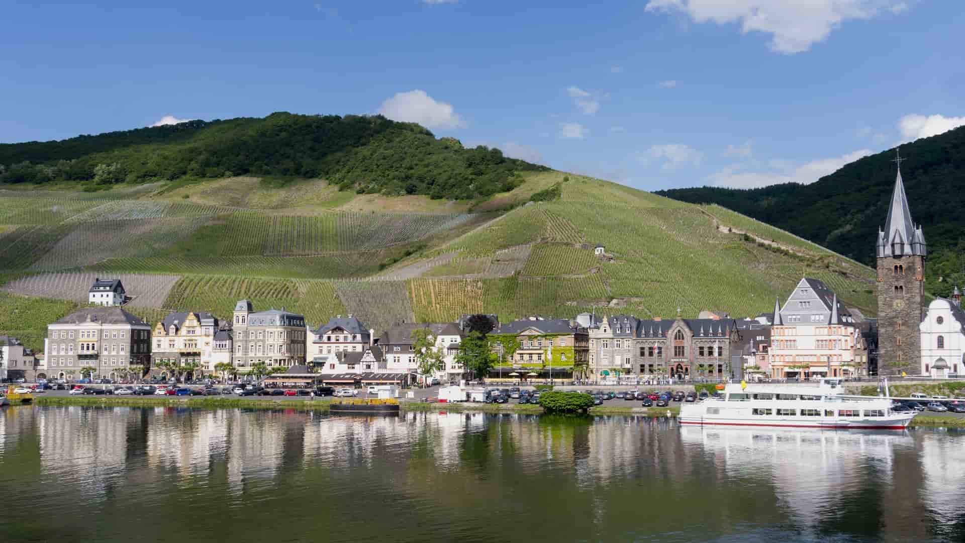 A beautiful landscape of Bernkastel-Kues, Germany, with a line of buildings and a ferry along the Moselle River. Terraced vineyards climb the steep hillside behind the town.