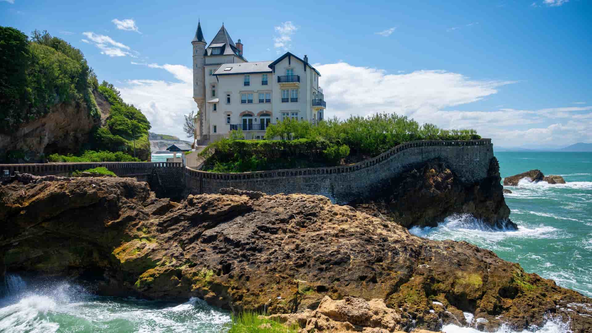 A white elegant building, Villa Belza, with a tower on a rocky outcrop overlooking the crashing waves of the Atlantic Ocean in Biarritz, France, on a sunny day.