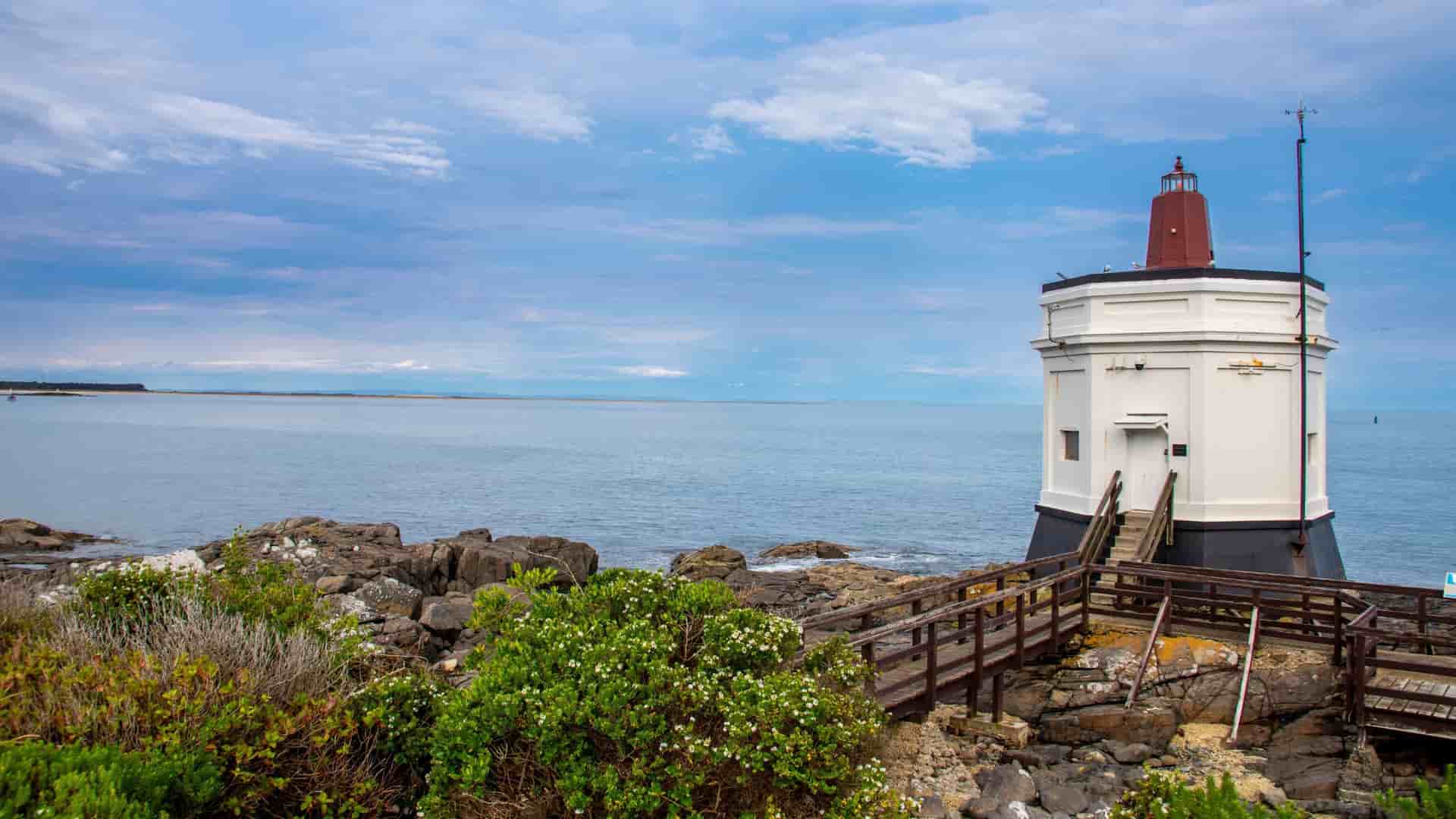 A scenic view of the iconic Bluff Lighthouse, a white and red structure with a wooden walkway, standing on a rocky shoreline overlooking the vast blue ocean under a cloudy sky.