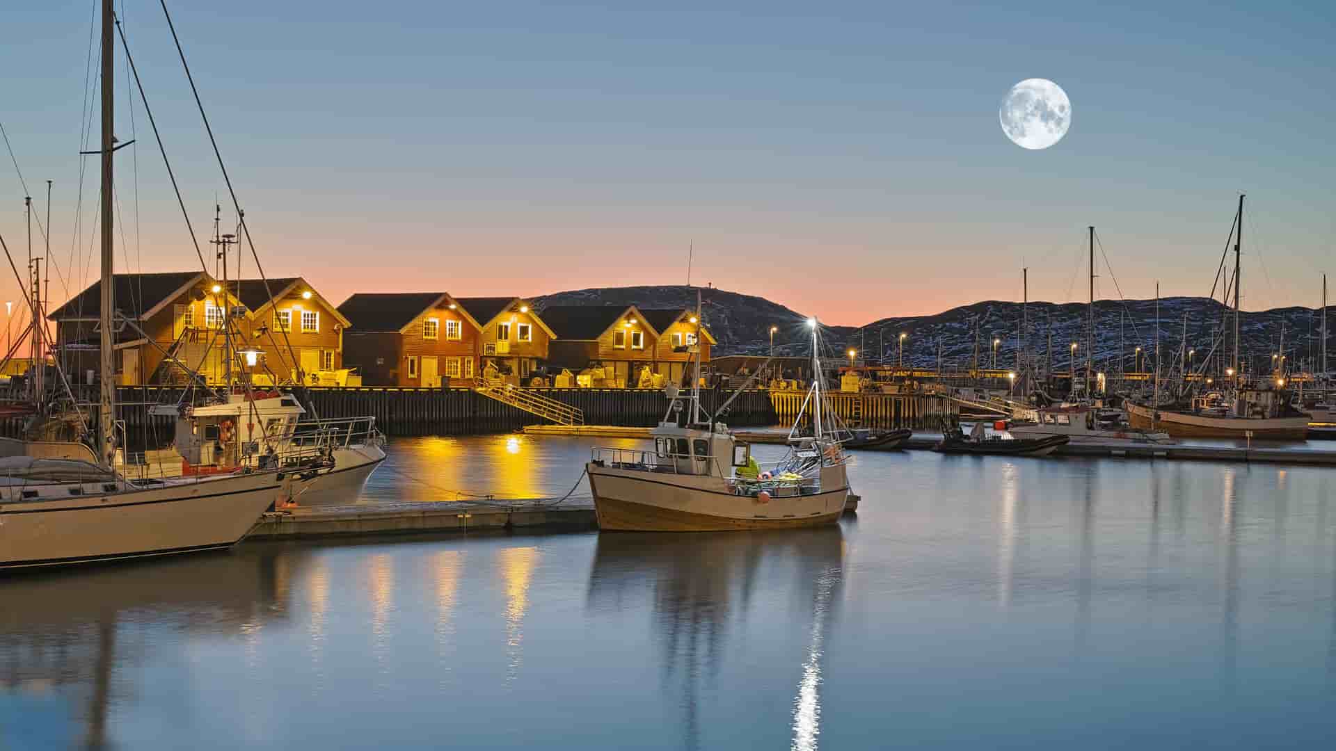 A scenic nighttime view of the harbor in Bodø, Norway, with boats and illuminated waterfront buildings under a full moon and a vibrant twilight sky.