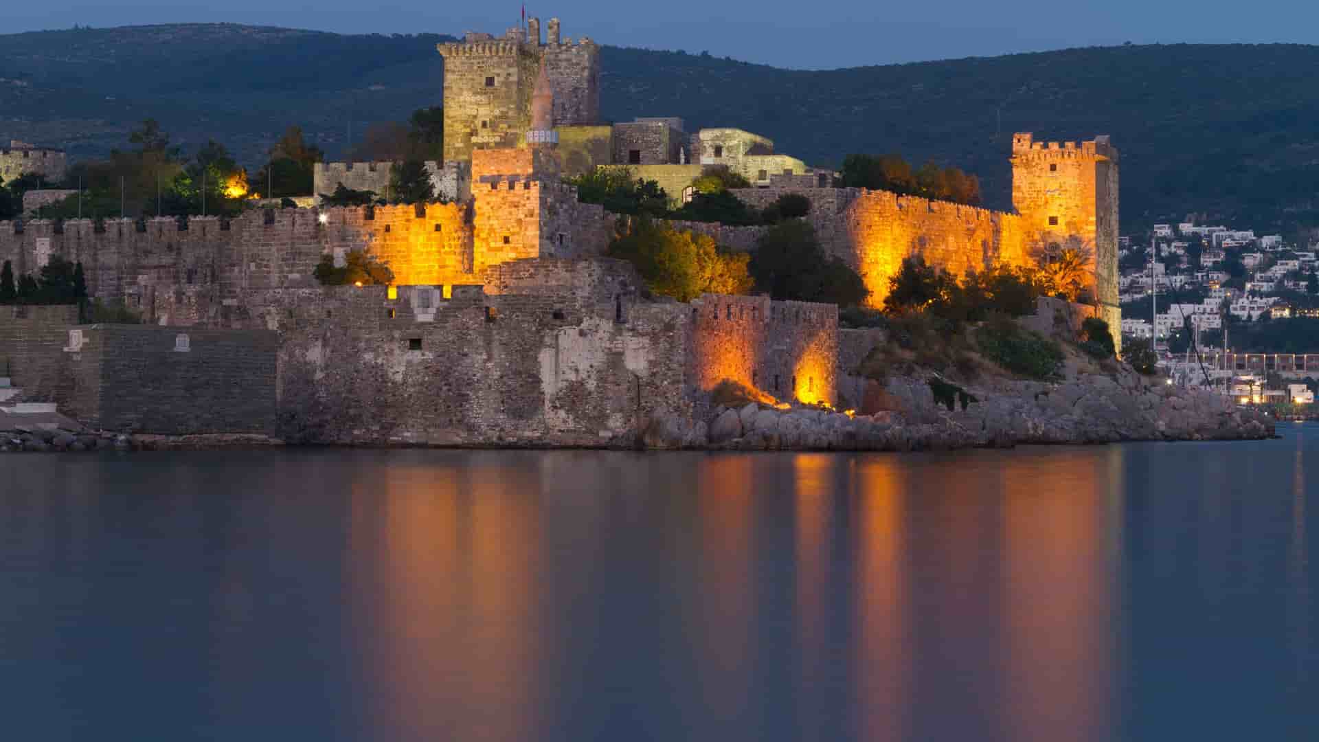 A dramatic dusk shot of the historic Bodrum Castle, also known as the Castle of St. Peter, illuminated at night, with its reflection visible in the calm waters of the harbor in Turkey.