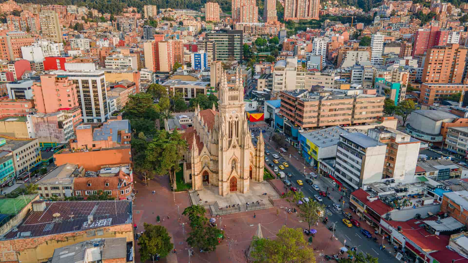 An aerial view of Bogota, Colombia, centered on the historic Church of Our Lady of Lourdes, a Gothic-style cathedral with twin spires. The church is surrounded by a large public square and a busy street filled with traffic. In the background, the city's densely packed buildings stretch up a green, forested hillside.