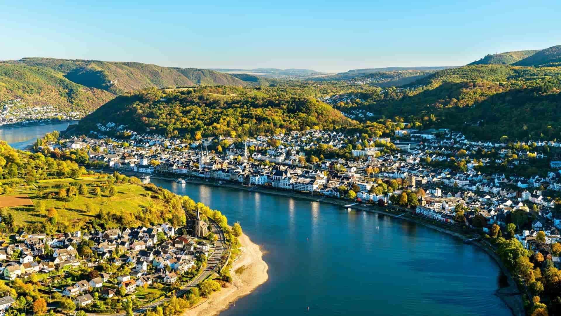 An aerial view of Boppard, a picturesque town along the Rhine River in Germany, nestled in a valley surrounded by vibrant green, forested hills and vineyards.