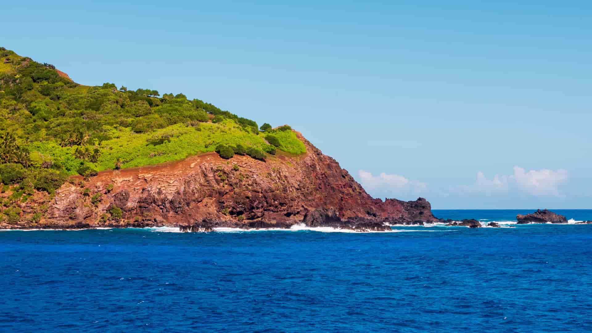 A photograph of Bounty Bay on Pitcairn Island, a British Overseas Territory in the South Pacific. The small bay is famous for being the final resting place of the HMS Bounty, which was burned by the mutineers who settled the island. The image shows the steep, rocky coastline of the island plunging down to the turquoise ocean, with a small landing area or dock visible at the edge of the bay. The island's slopes are covered in lush green tropical vegetation.