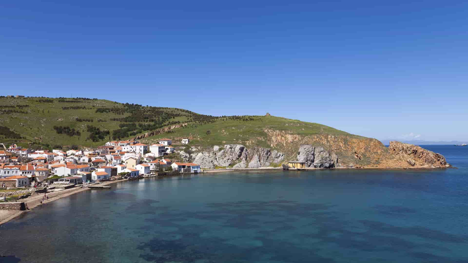 A beautiful scenic view of the coastal town of Bozcaada, Turkey, with its charming white houses nestled at the foot of a grassy hill and dramatic cliffs overlooking a clear bay.