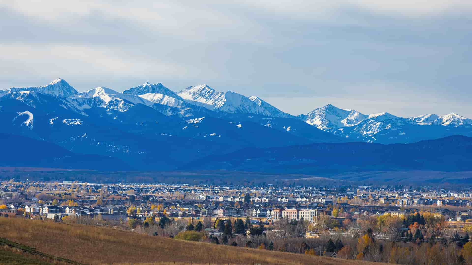 A panoramic view of Bozeman, Montana, with the snow-capped peaks of the Bridger Mountains dominating the background. The town fills the midground, with houses and buildings surrounded by trees displaying yellow and orange autumn foliage. A brown, grassy hill is in the foreground under a partially cloudy blue sky.