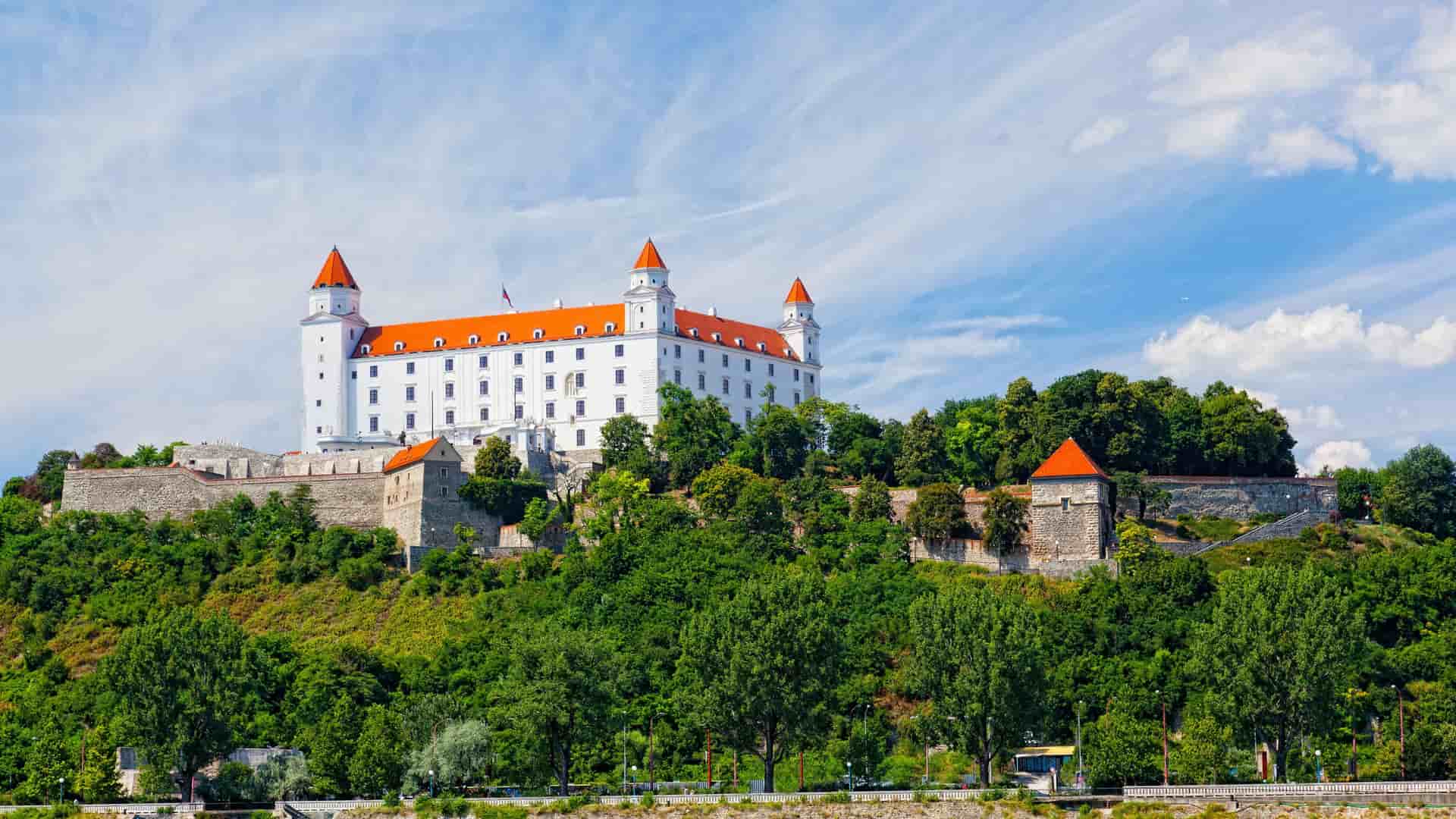 A scenic view of the white Bratislava Castle, a famous Slovak landmark, with its four distinctive red-tiled towers, perched on a lush, green hill overlooking the Danube River on a sunny day.