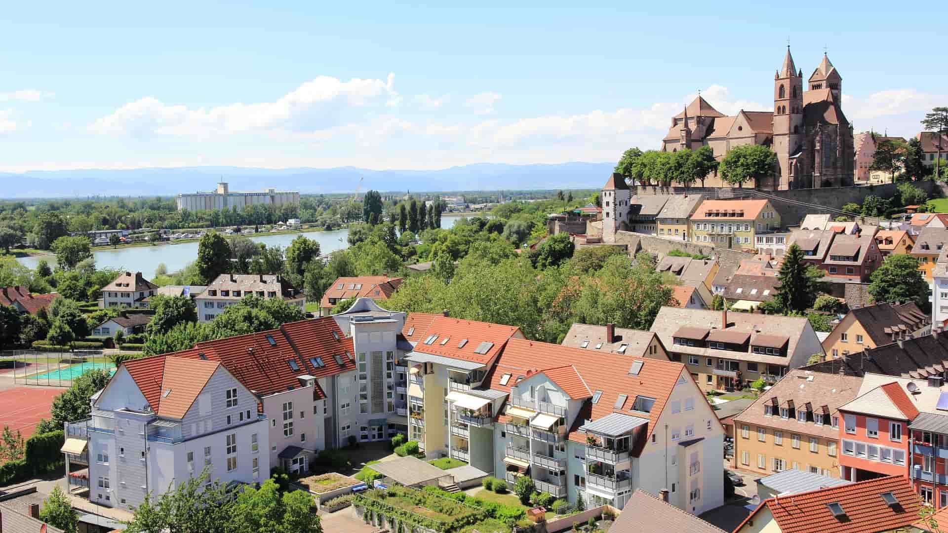 A scenic panorama of Breisach, Germany, showcasing the medieval St. Stephen's Cathedral prominently on a hill overlooking the colorful rooftops of the town, with the Rhine River in the background.