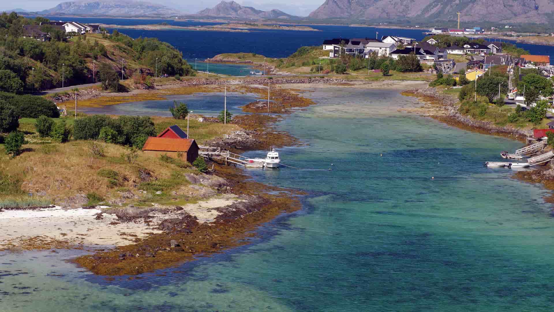 A stunning coastal landscape of Brønnøysund, Norway, with a small boathouse on the shore of a clear bay, surrounded by a charming village and rugged mountains in the distance.