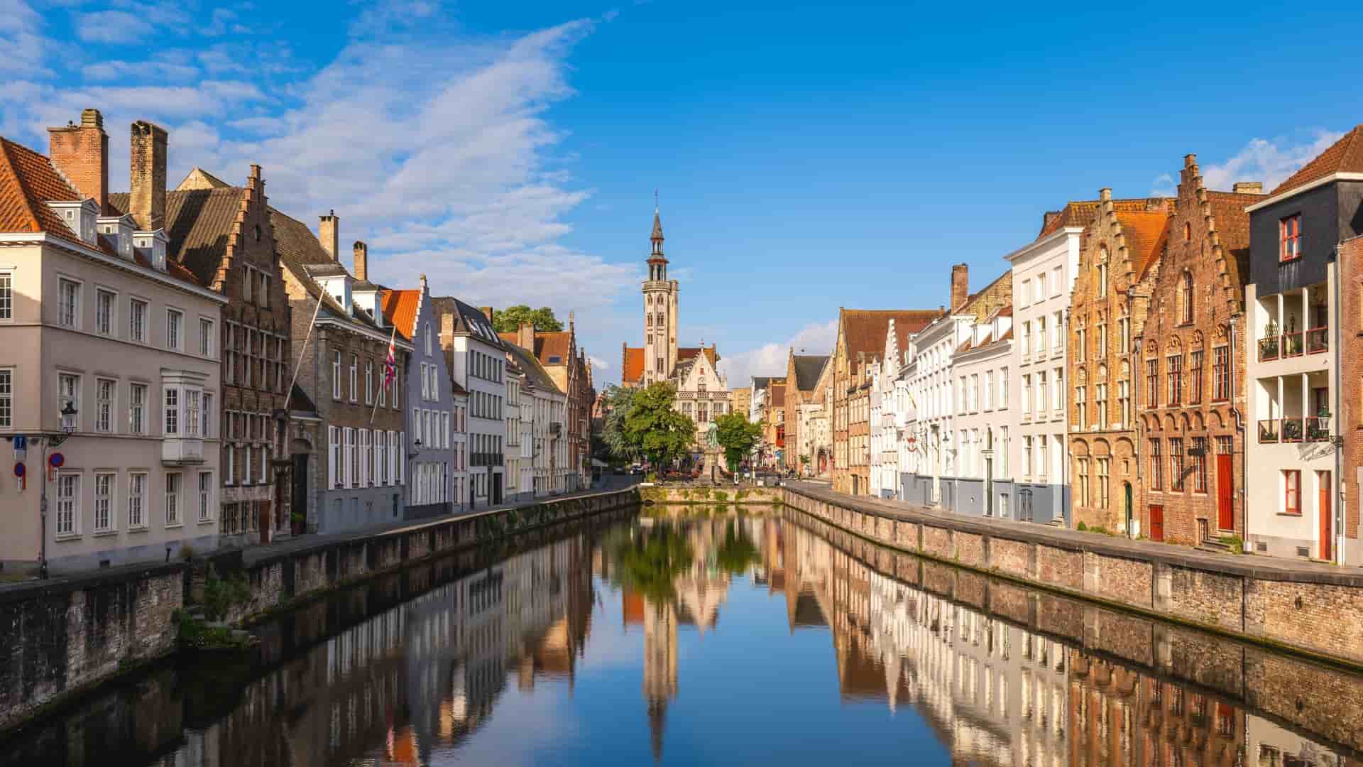A scenic canal in Bruges, Belgium, with colorful medieval buildings and their reflections perfectly captured in the calm water, under a bright blue sky with scattered clouds.