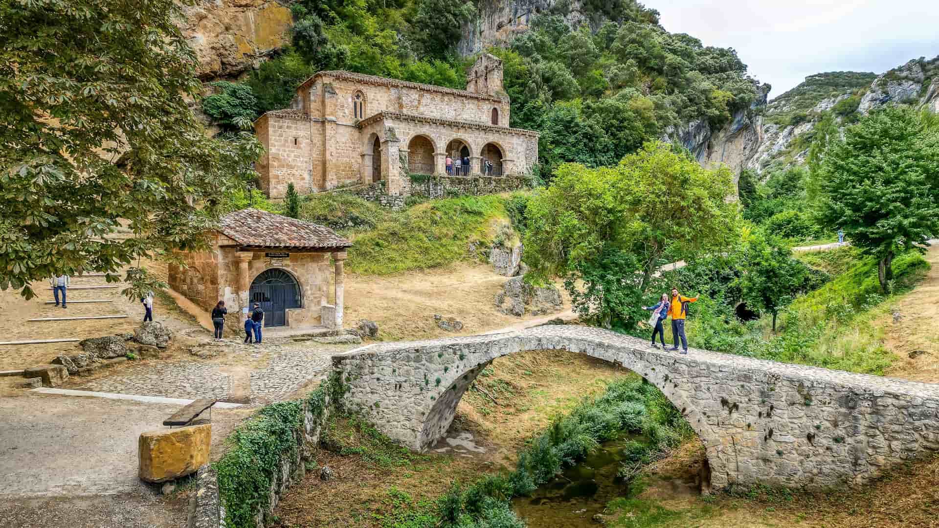 A view of the historic Hermitage of San Bernabé near Burgos, Spain, nestled into a green, rocky hillside. In the foreground, a small, ancient stone arch bridge crosses a stream. People are visible on the bridge and on the path leading to the hermitage, which is surrounded by lush trees and a smaller stone building.