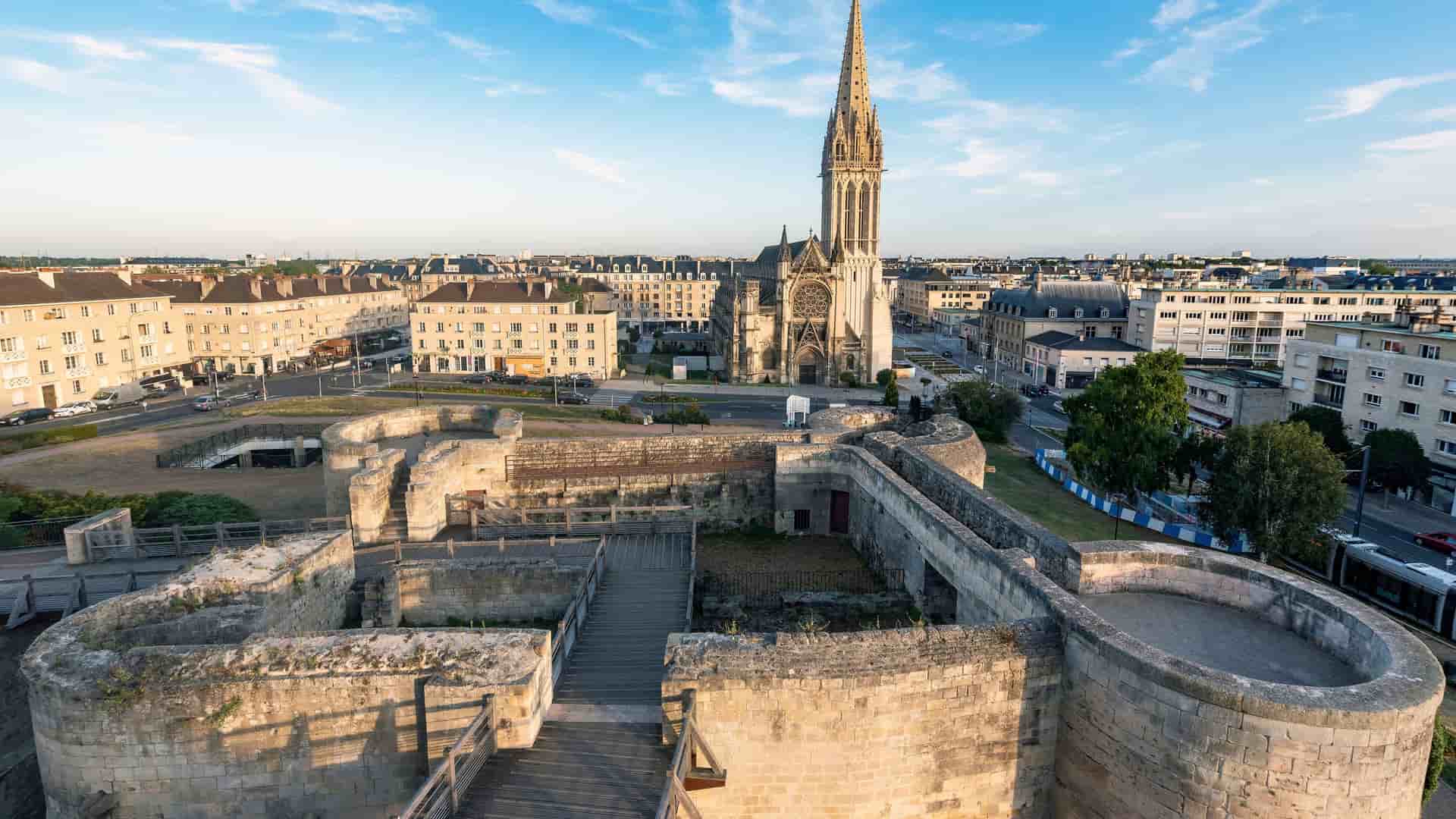 An elevated view of the historic city of Caen, France, at sunset. In the foreground are the extensive stone ruins of a medieval castle, with a wooden walkway for visitors. In the background, the ornate, towering spire of the Église Saint-Pierre rises above the surrounding urban landscape of modern and historic buildings under a clear blue sky.