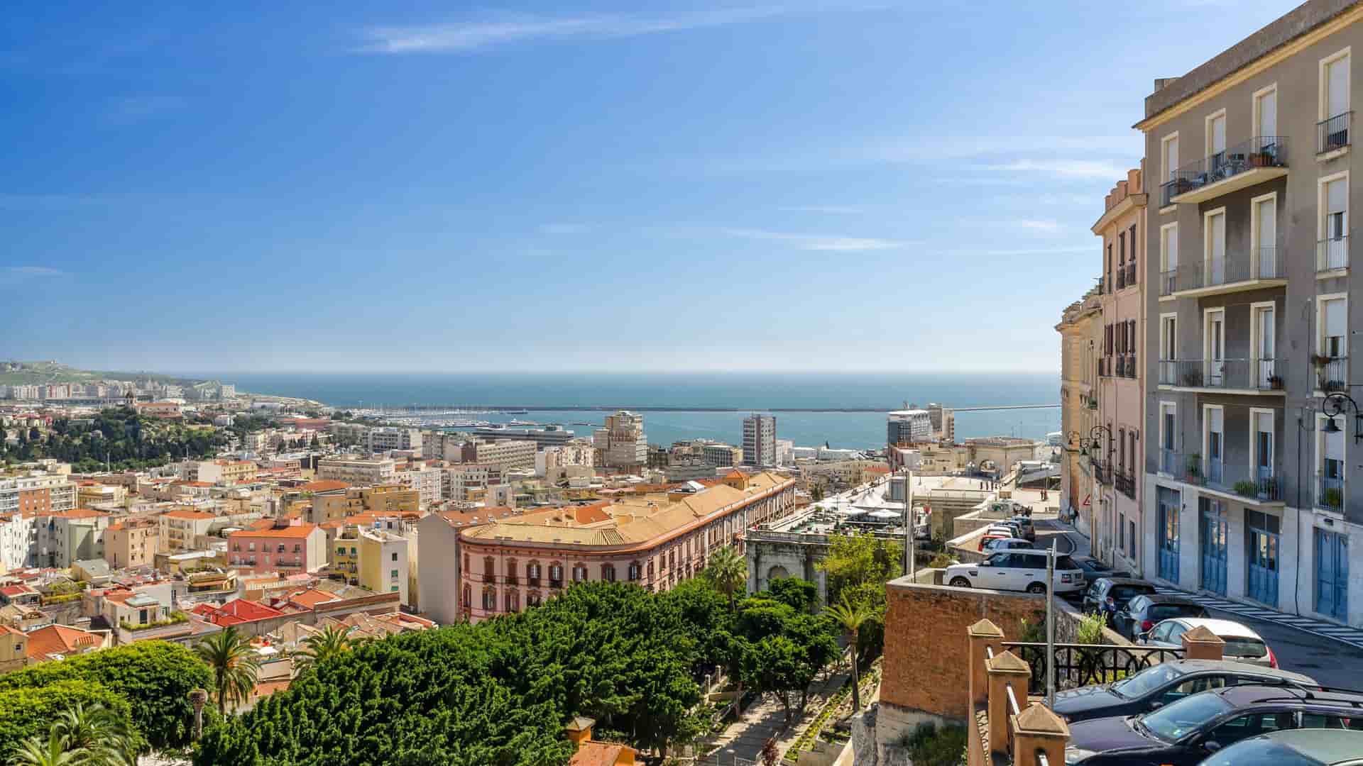 A high-angle view of the bustling city of Cagliari, Sardinia, Italy, with historic buildings and rooftops overlooking the Mediterranean Sea under a clear blue sky.
