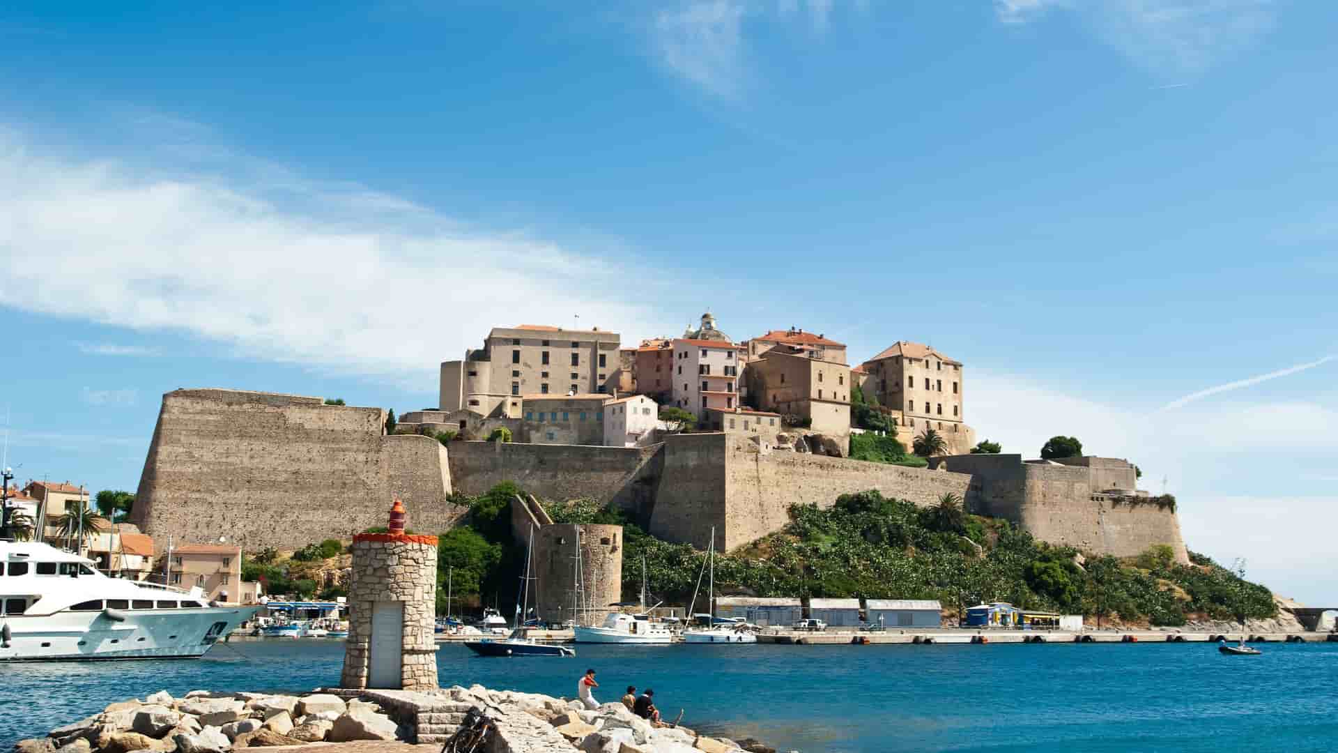 A stunning view of the historic Calvi Citadel on the Corsican coastline, with its fortified walls overlooking a vibrant harbor filled with boats and a clear blue sky.