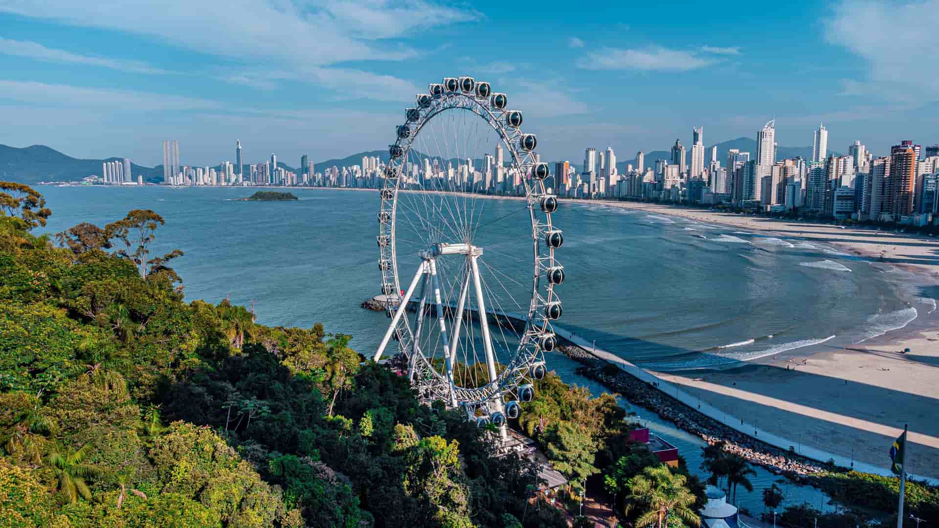 An aerial view of the Balneário Camboriú Ferris wheel, a landmark attraction on the Brazilian coastline, with the city's impressive skyline and sandy beach in the background.