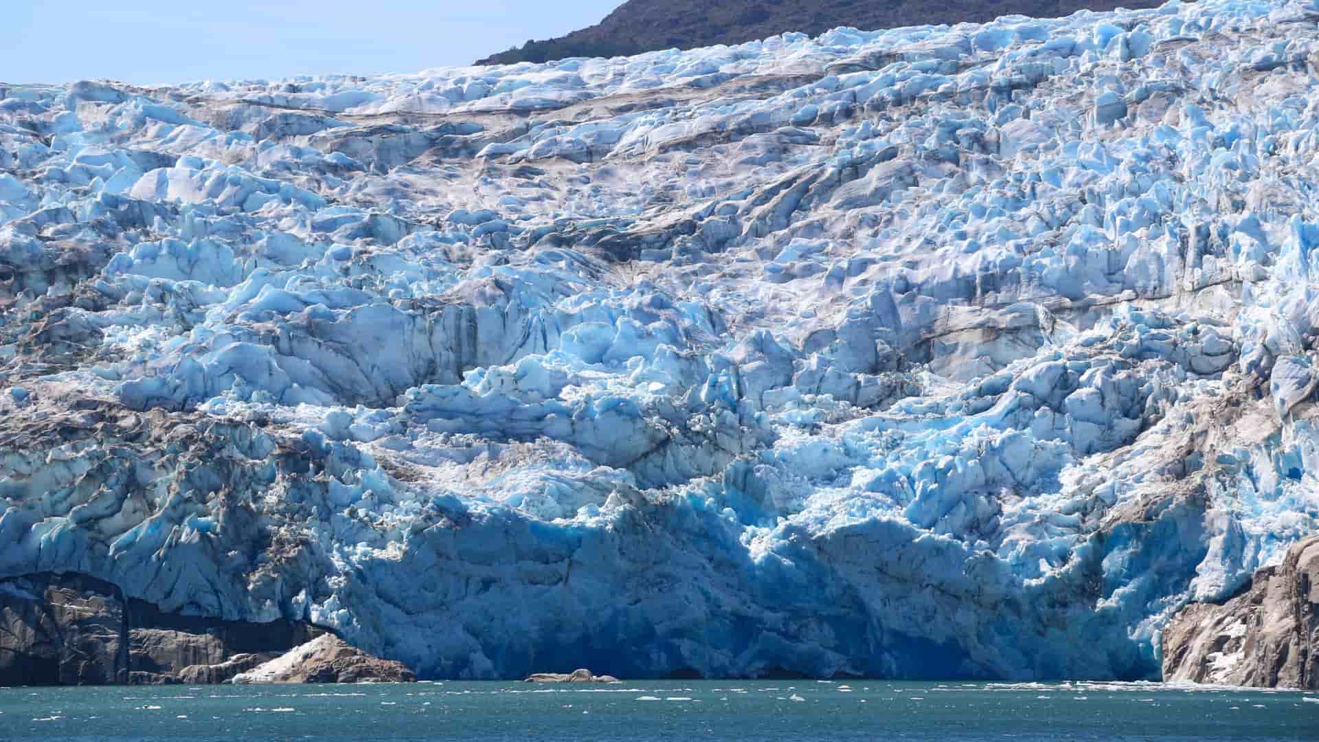 A massive, stunning blue and white glacier in the Sarmiento Channel, Patagonia, Chile, with its dramatic crevasses and ice formations towering over the deep blue water.