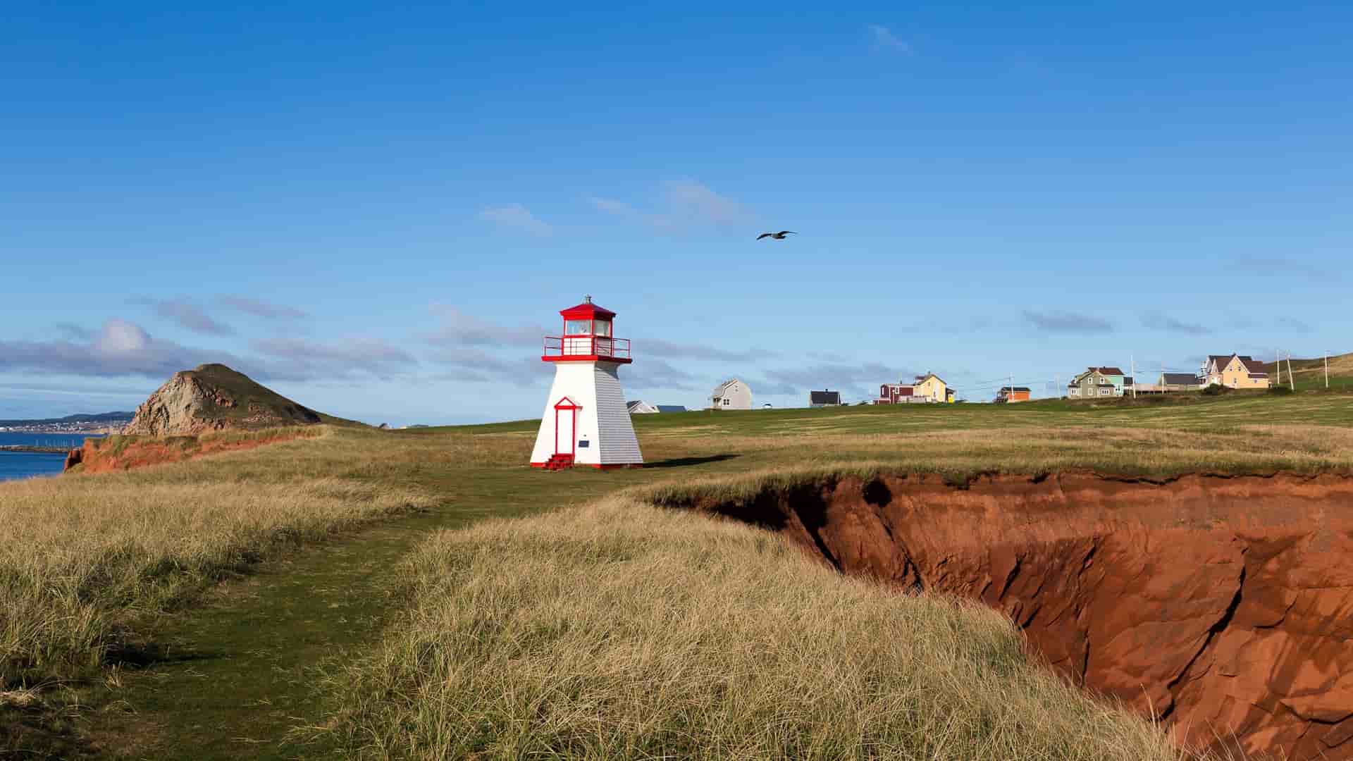 A charming white and red lighthouse, perched on a dramatic red cliff overlooking the ocean, with lush green grass and a few houses under a bright blue sky on Cap-aux-Meules, Quebec.