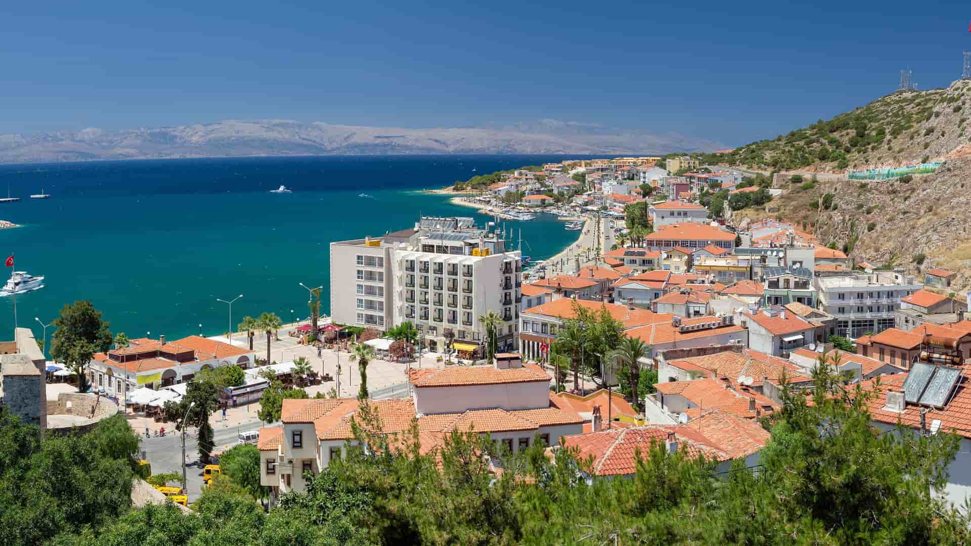 A high-angle view of the bustling coastal town of Cesme, Turkey, featuring a marina filled with boats, white buildings with red-tiled roofs, and the Aegean Sea.