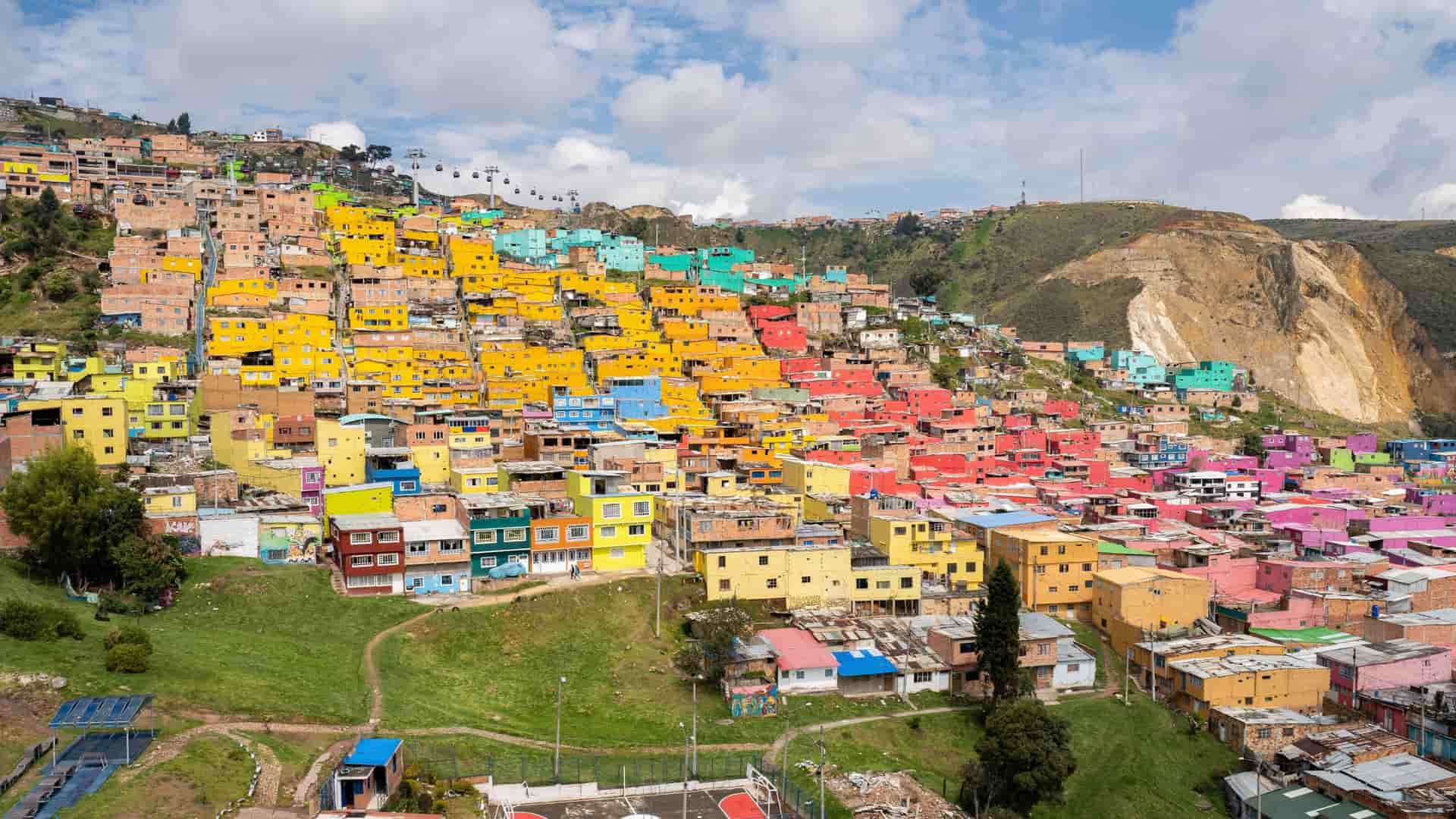 A colorful residential area of Ciudad Bolivar, Bogotá, with bright yellow, blue, and pink houses built on a hillside, with a cable car visible in the sky.
