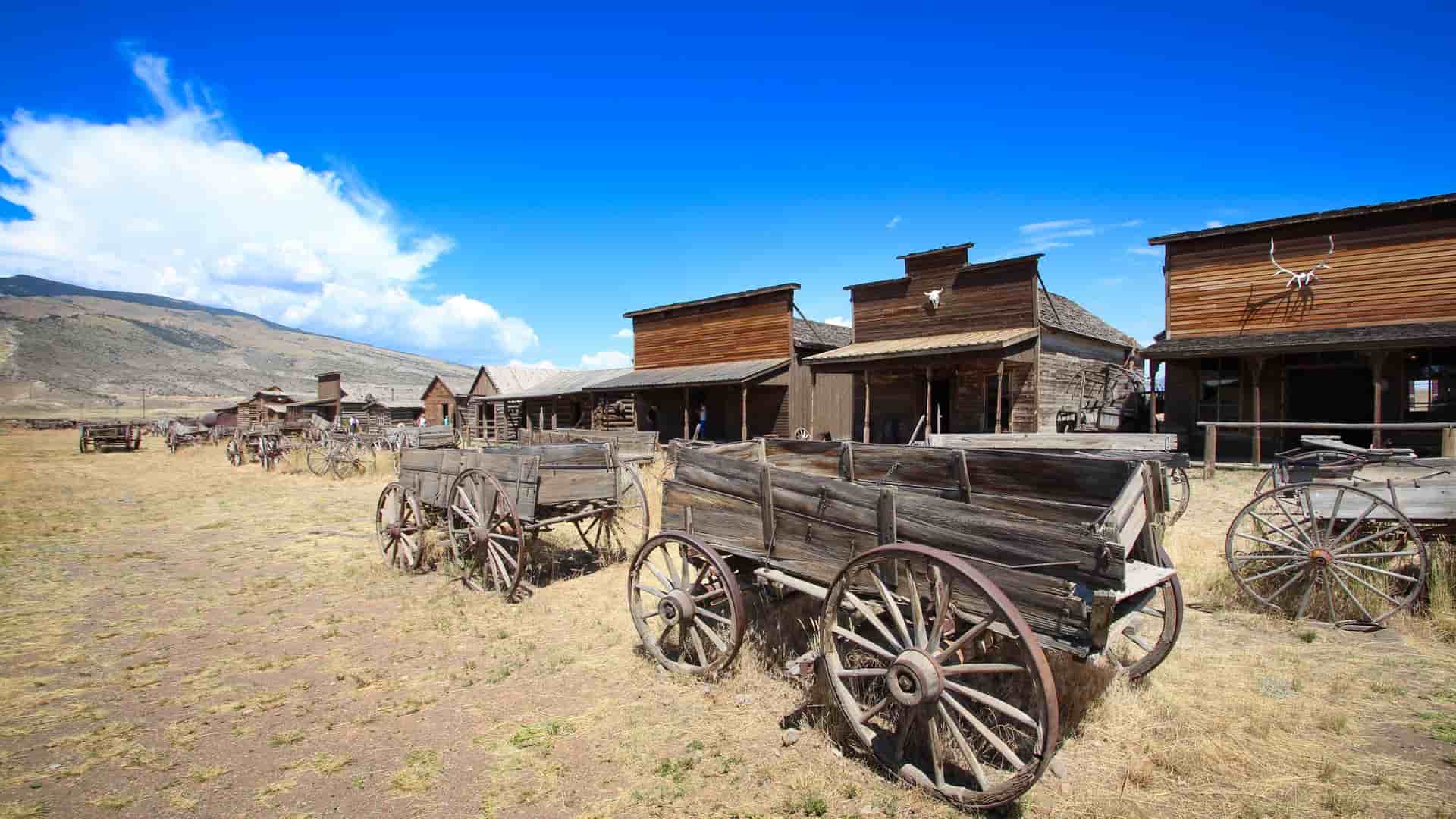 A historic wild west ghost town with weathered wooden buildings and old, rustic covered wagons on display under a bright blue sky with white clouds in Cody, Wyoming.