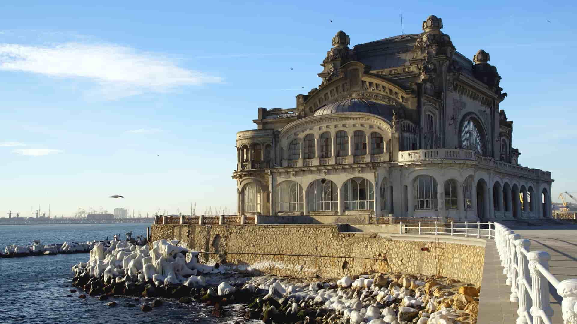 A grand, abandoned Art Nouveau casino building on the waterfront of Constanța, Romania, with icy rocks in the foreground and the Black Sea in the background.