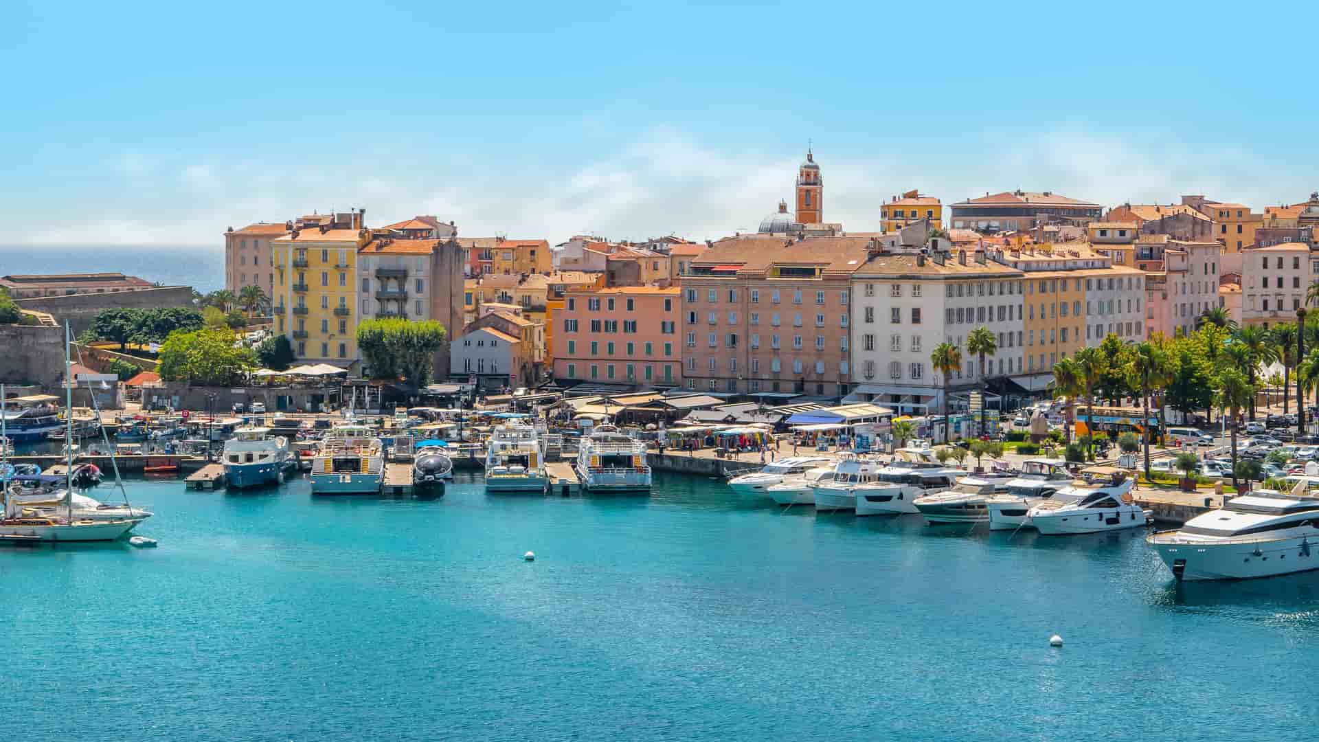 A beautiful view of the bustling harbor in Ajaccio, Corsica, with colorful historic buildings lining the port and numerous yachts docked in the marina.