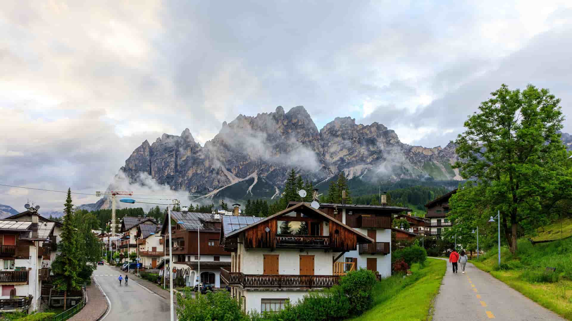 "A picturesque village scene in Cortina d'Ampezzo, Italy, with charming alpine buildings and a couple of people walking on a path, set against the dramatic, rocky peaks of the Dolomite mountains under a cloudy sky. "