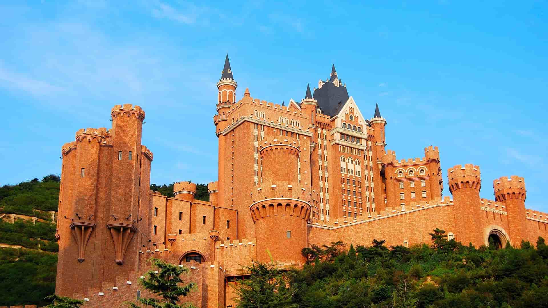 A majestic modern castle, the Dalian Castle Hotel, with its red brick facade and multiple turrets, stands on a hillside in Dalian, China.