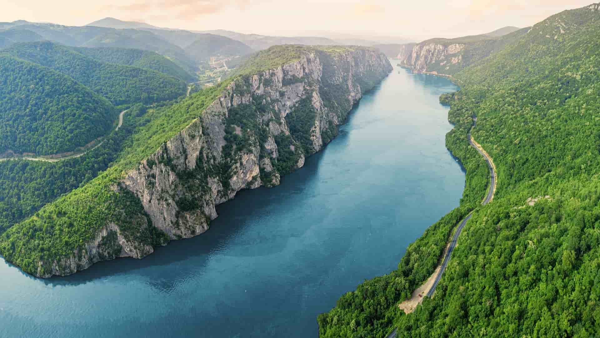 An aerial view of the wide Danube River flowing through the dramatic Iron Gates gorge, flanked by a massive cliff and lush green forests.