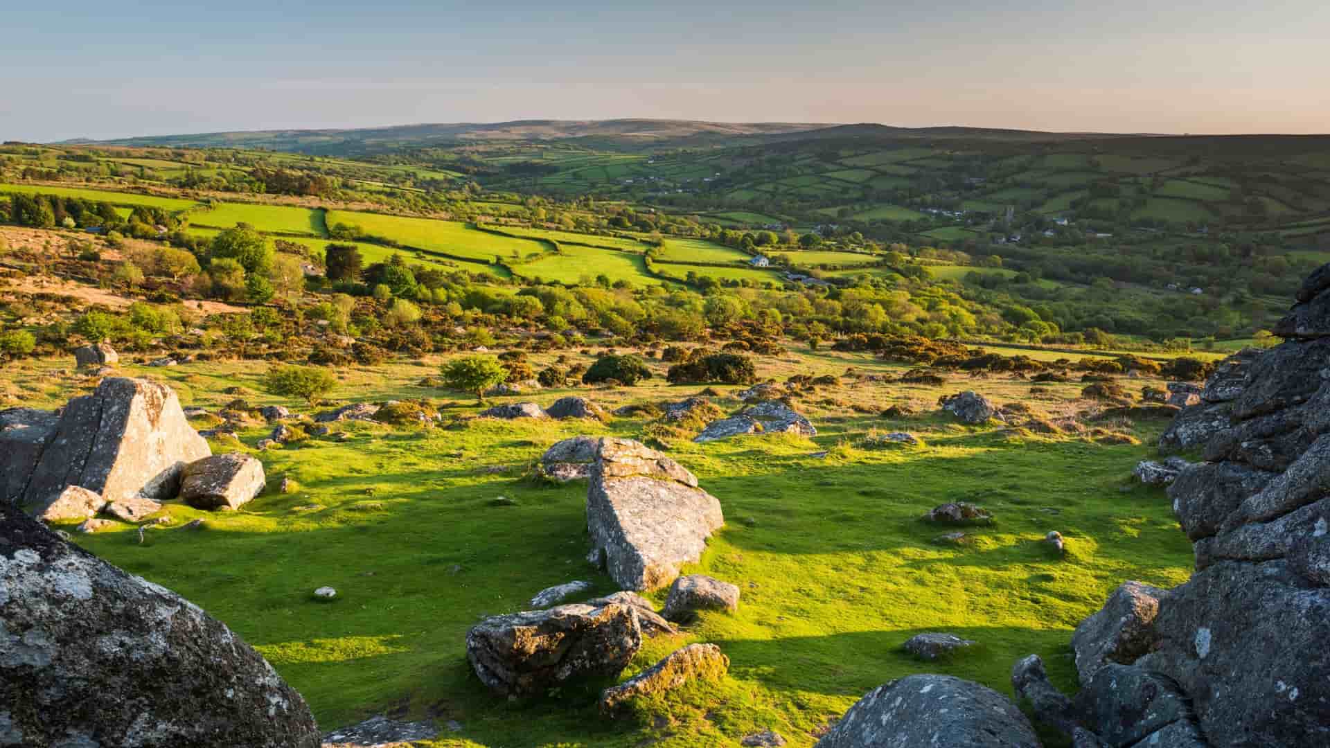 Granite tors and lush green landscape in Dartmoor National Park, Devon, UK. View of farmlands and rolling hills at sunset.