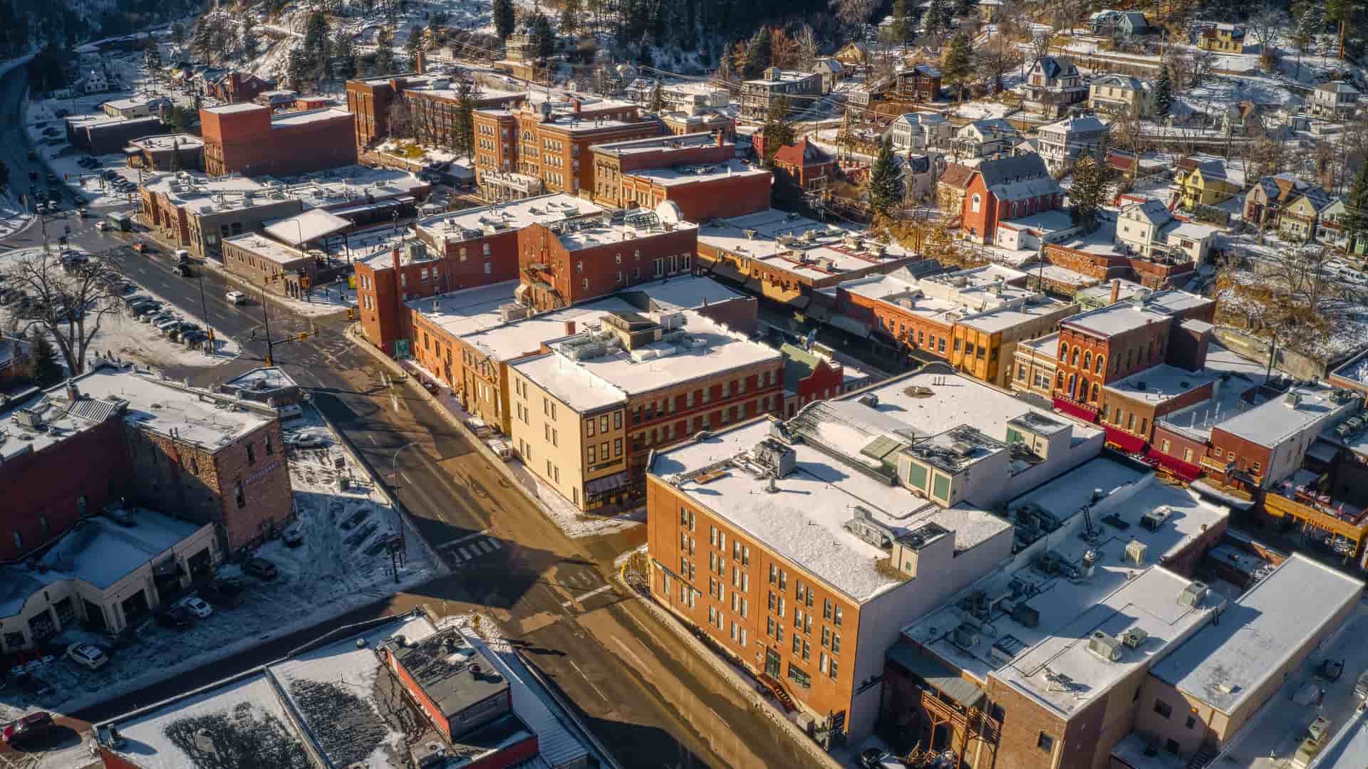 "An aerial view of the historic town of Deadwood, South Dakota, with its brick buildings and rooftops lightly covered in a fresh layer of snow, nestled in a valley among the pine-covered Black Hills.  "