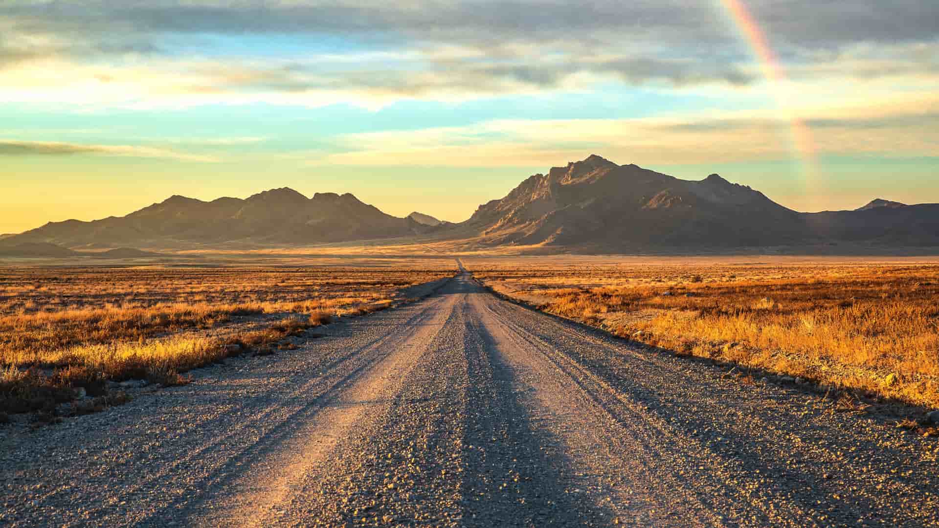 A long gravel road disappearing into the distance through a golden-lit desert landscape with rugged mountains and a faint rainbow at sunrise.
