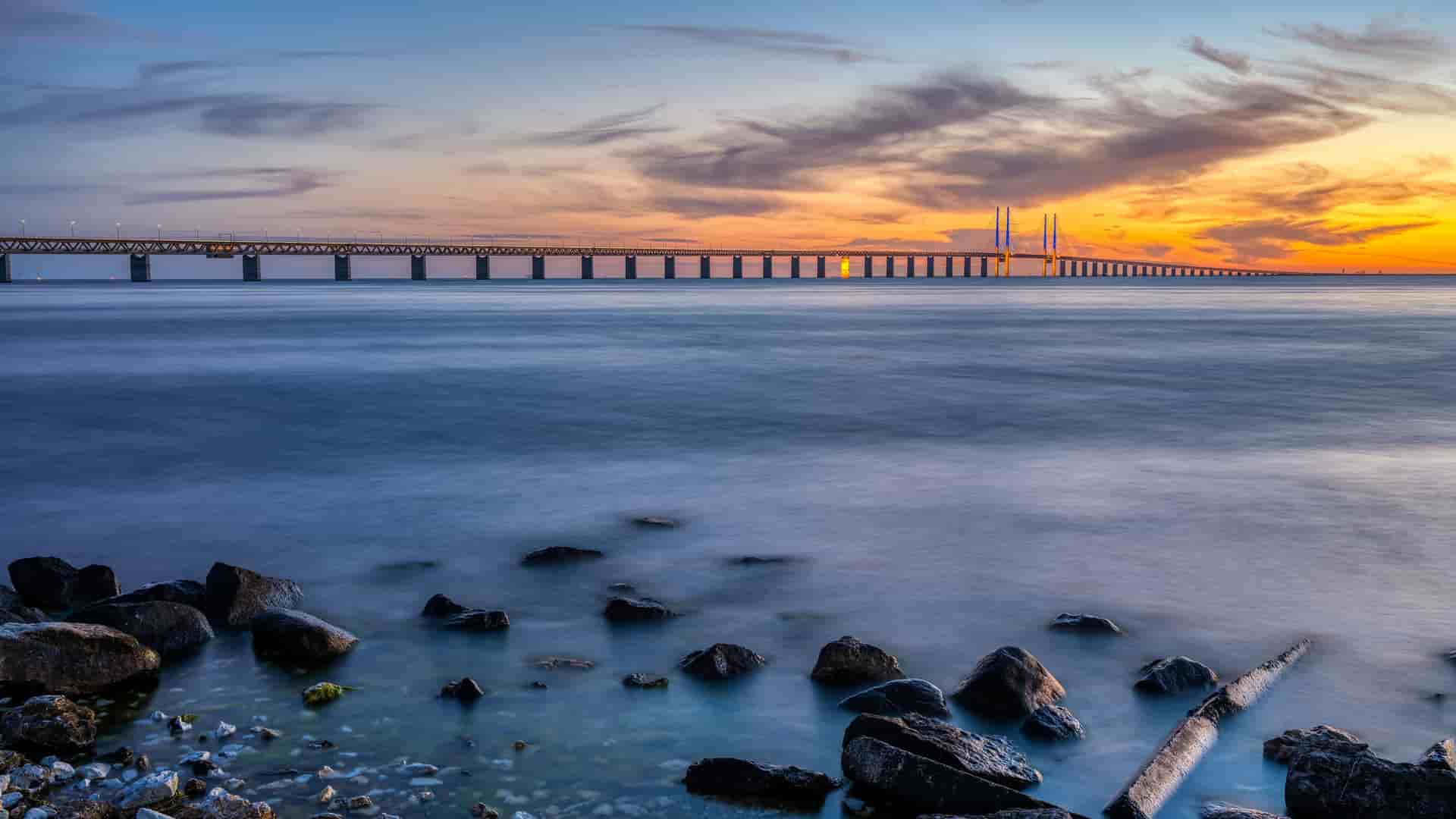 The iconic Øresund Bridge connecting Denmark and Sweden, captured in a long exposure shot at sunset with beautiful colors reflecting on the water.
