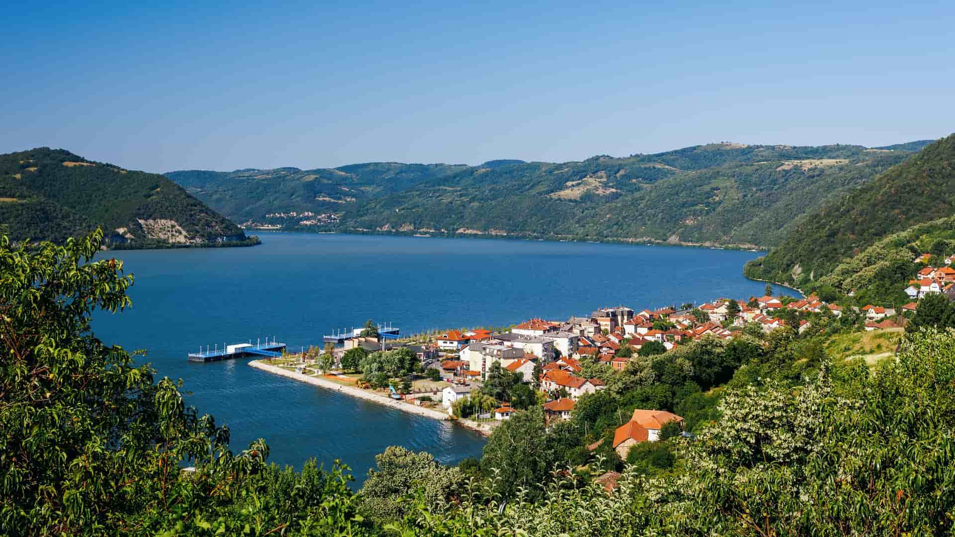 The picturesque town of Donji Milanovac, Serbia, nestled on the wide banks of the Danube River, surrounded by the lush, green hills of Đerdap National Park.