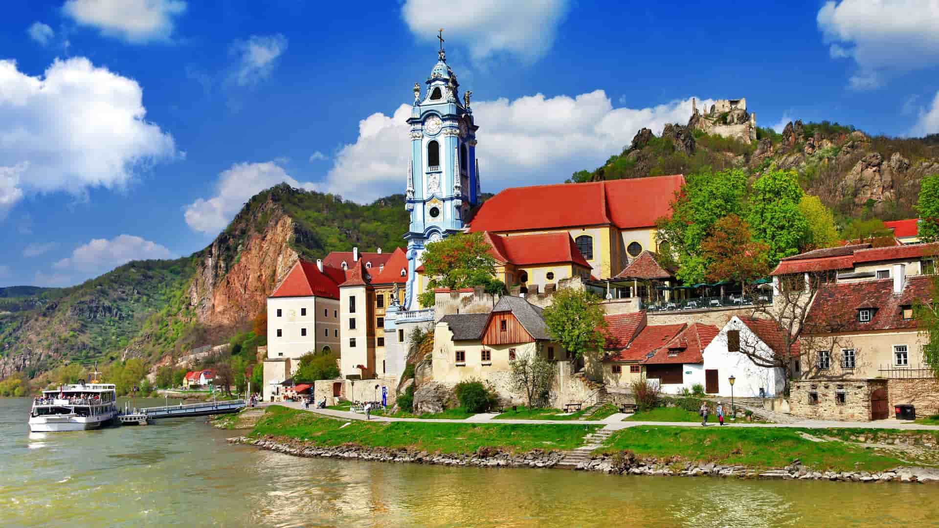 A view of the historic town of Dürnstein, Austria, on the Danube River, featuring a picturesque blue and white church tower, red-roofed buildings, and the Kuenringerburg castle ruins on the hillside.