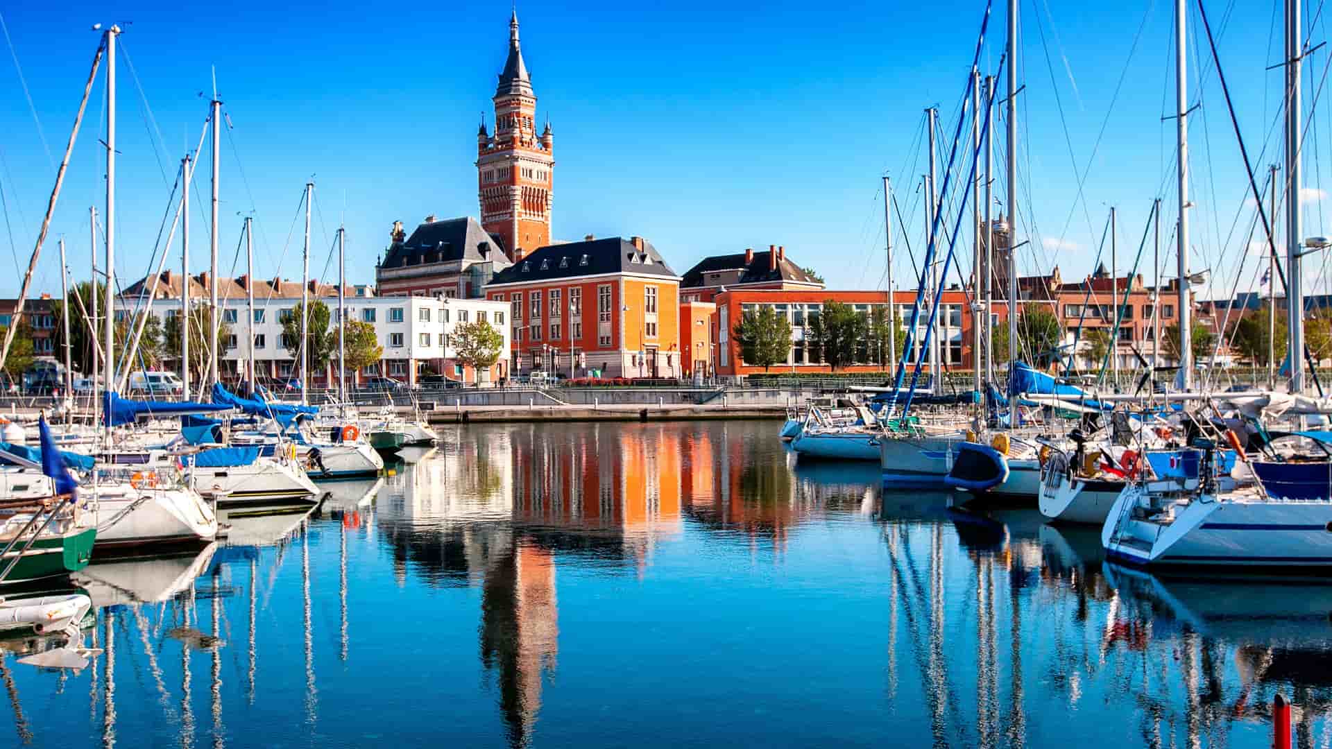 A picturesque view of the harbor in Dunkirk, France, with a marina full of sailboats and a historic brick building with a prominent clock tower reflecting on the calm water.
