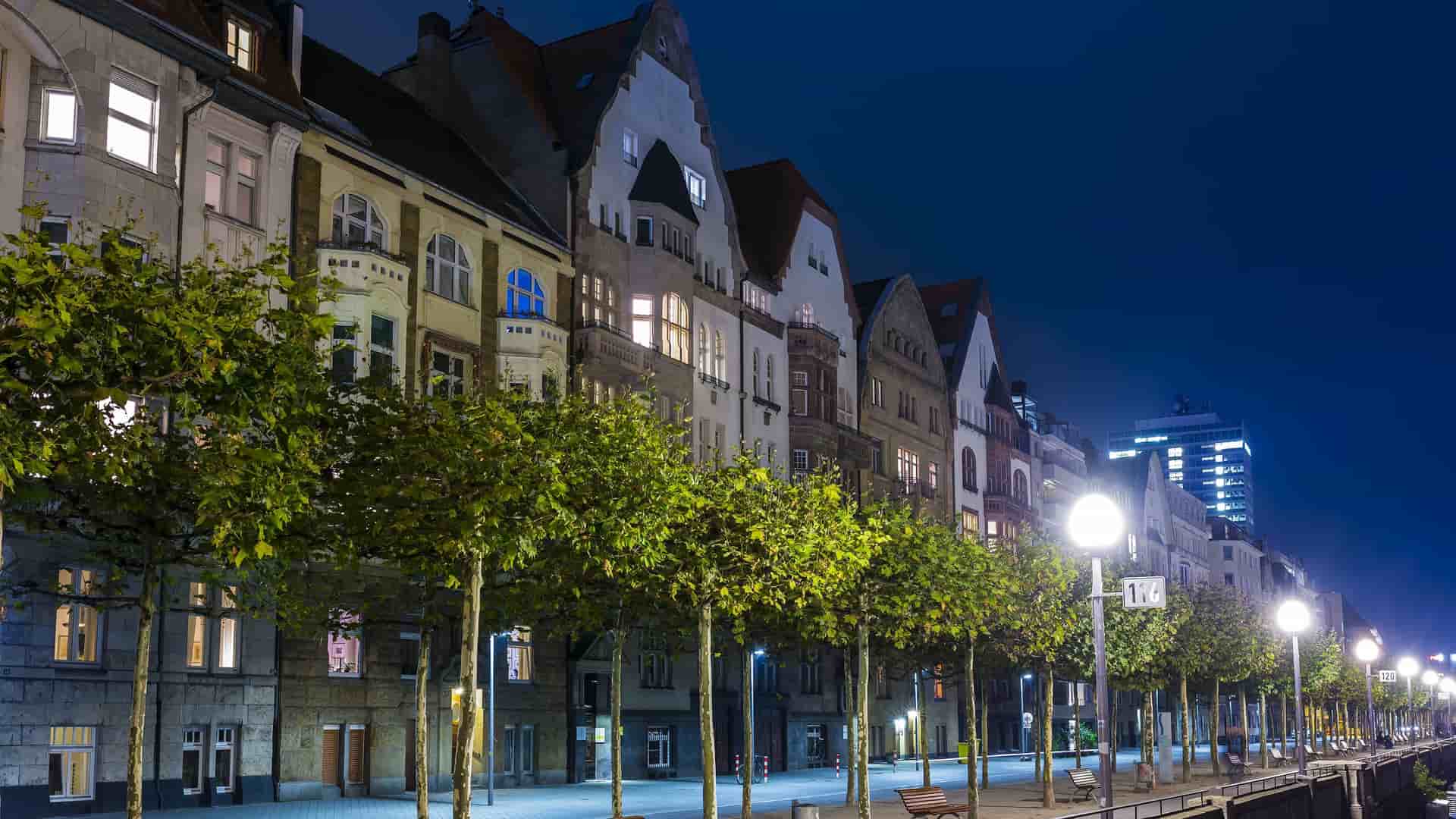 A serene nighttime shot of a street in Düsseldorf, Germany, with a line of trees and old buildings with glowing windows lining the street under a deep blue sky.