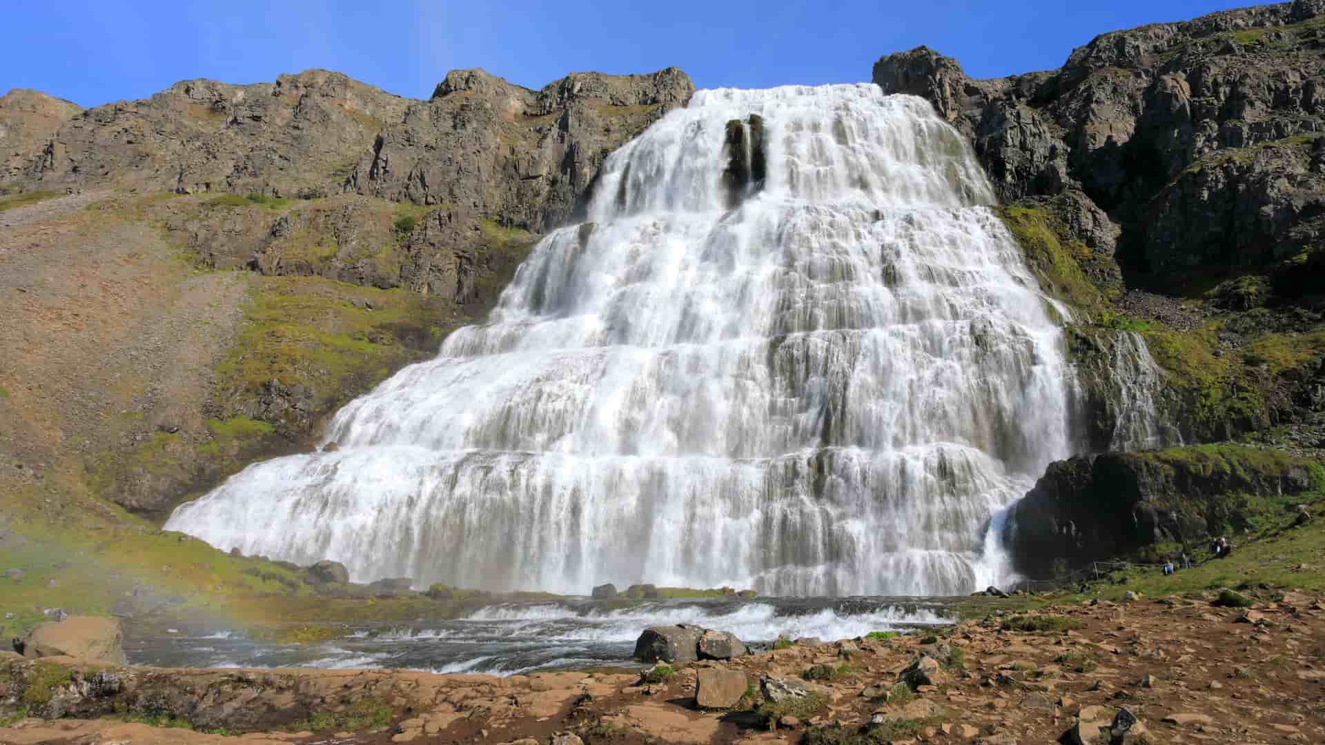 A majestic view of the powerful Dynjandi waterfall in the Westfjords of Iceland, cascading down a series of tiered cliffs with a small rainbow visible at its base.