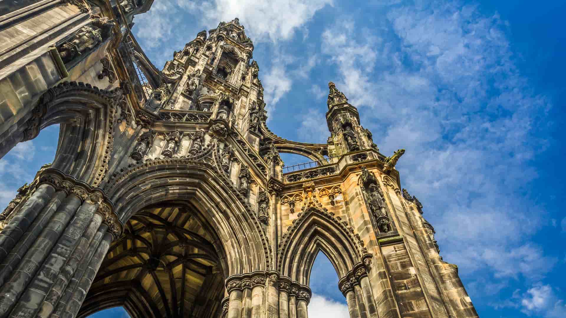 A dramatic low-angle shot of the Sir Walter Scott Monument in Edinburgh, Scotland, showcasing the intricate Gothic architecture of the spire against a brilliant blue sky with wispy clouds.