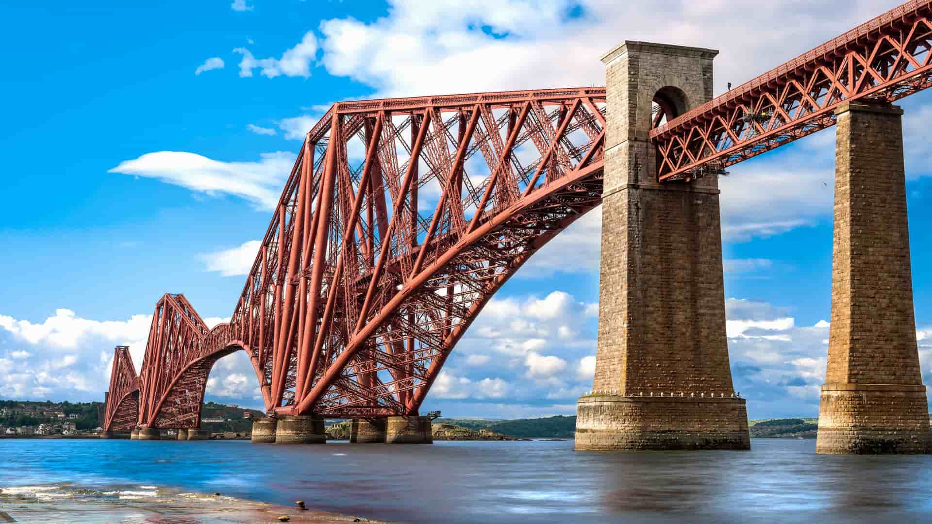A low-angle shot of the iconic Forth Bridge near Edinburgh, Scotland, showcasing its distinctive red cantilever design and massive stone pillars stretching across the water under a bright, cloudy sky.