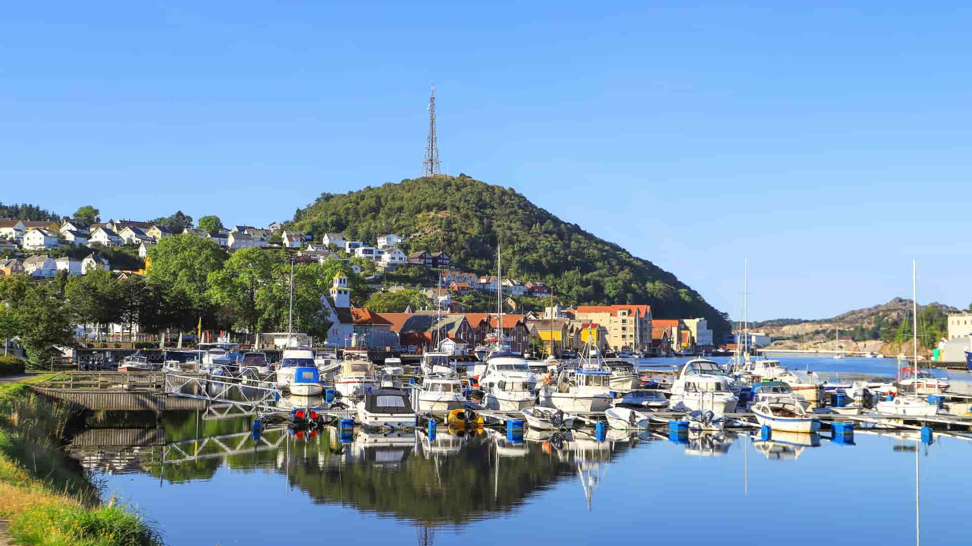 A serene view of the Egersund harbor in Norway, with a marina full of boats reflecting on the calm water, surrounded by colorful buildings and a steep, green hill.