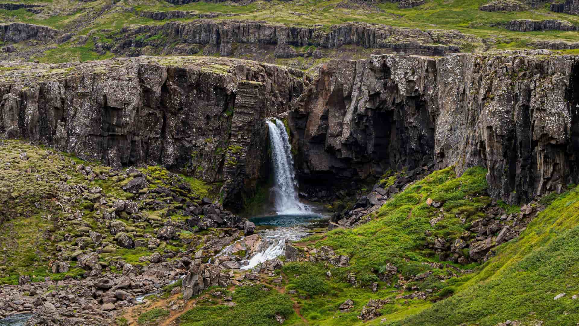 "A powerful waterfall cascading over a cliff face into a small turquoise pool, surrounded by rugged, rocky cliffs and vibrant green moss in the scenic landscape near Egilsstaðir, Iceland.  "