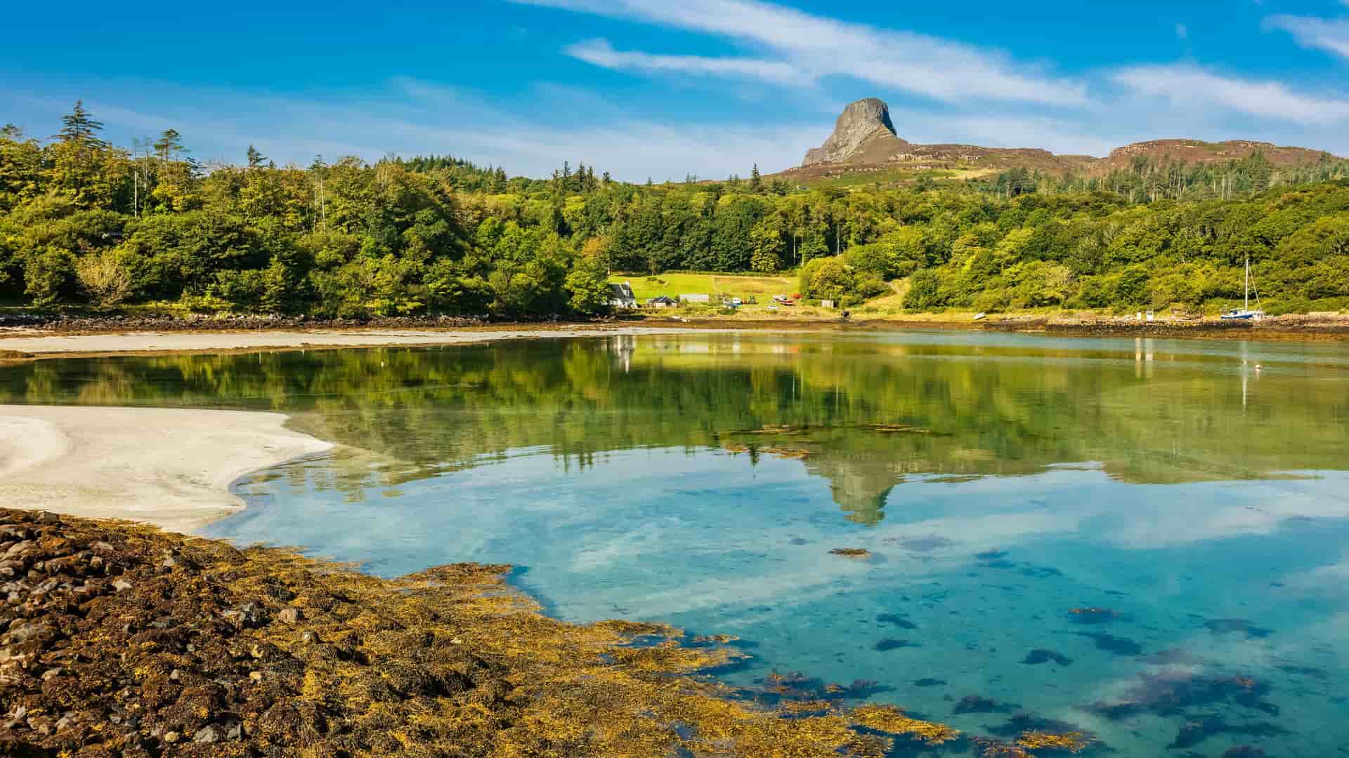 A scenic view of the Isle of Eigg, Scotland, showcasing a beautiful turquoise bay with a sandy beach and a distinctive hill, An Sgurr, in the distance, surrounded by lush green forests.