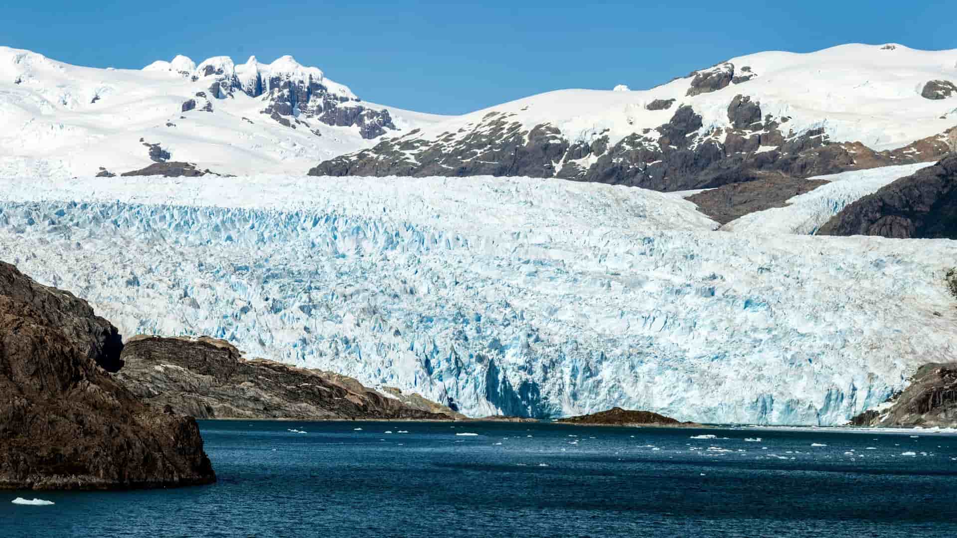A breathtaking view of El Brujo Glacier in Chile, with its massive, textured blue ice wall flowing from snow-capped mountains into a deep blue body of water with floating ice chunks.
