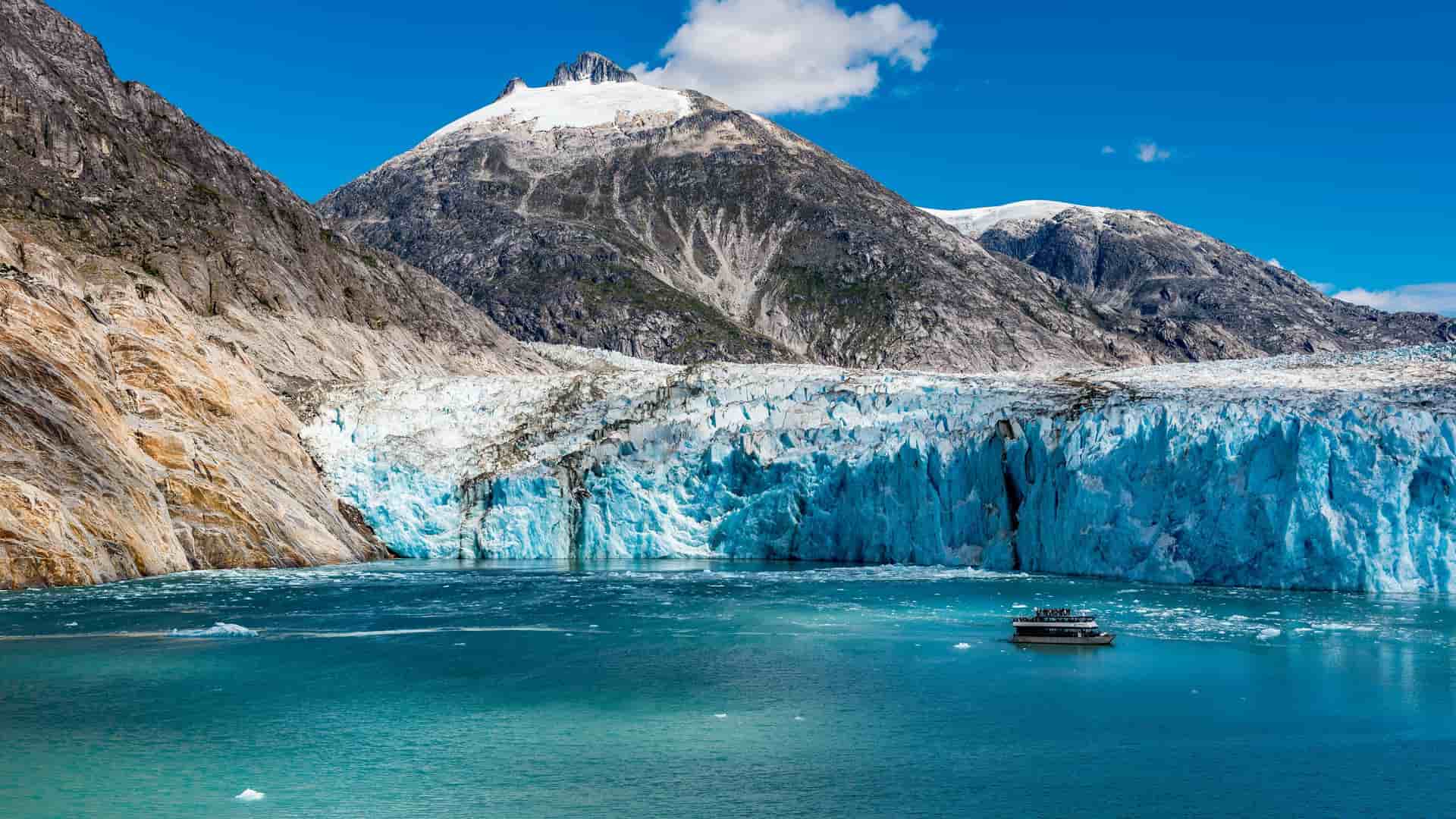 A tour boat navigates the turquoise water in front of the massive Dawes Glacier, with its textured blue face, and a snow-covered mountain in the background at Endicott Arm, Alaska.