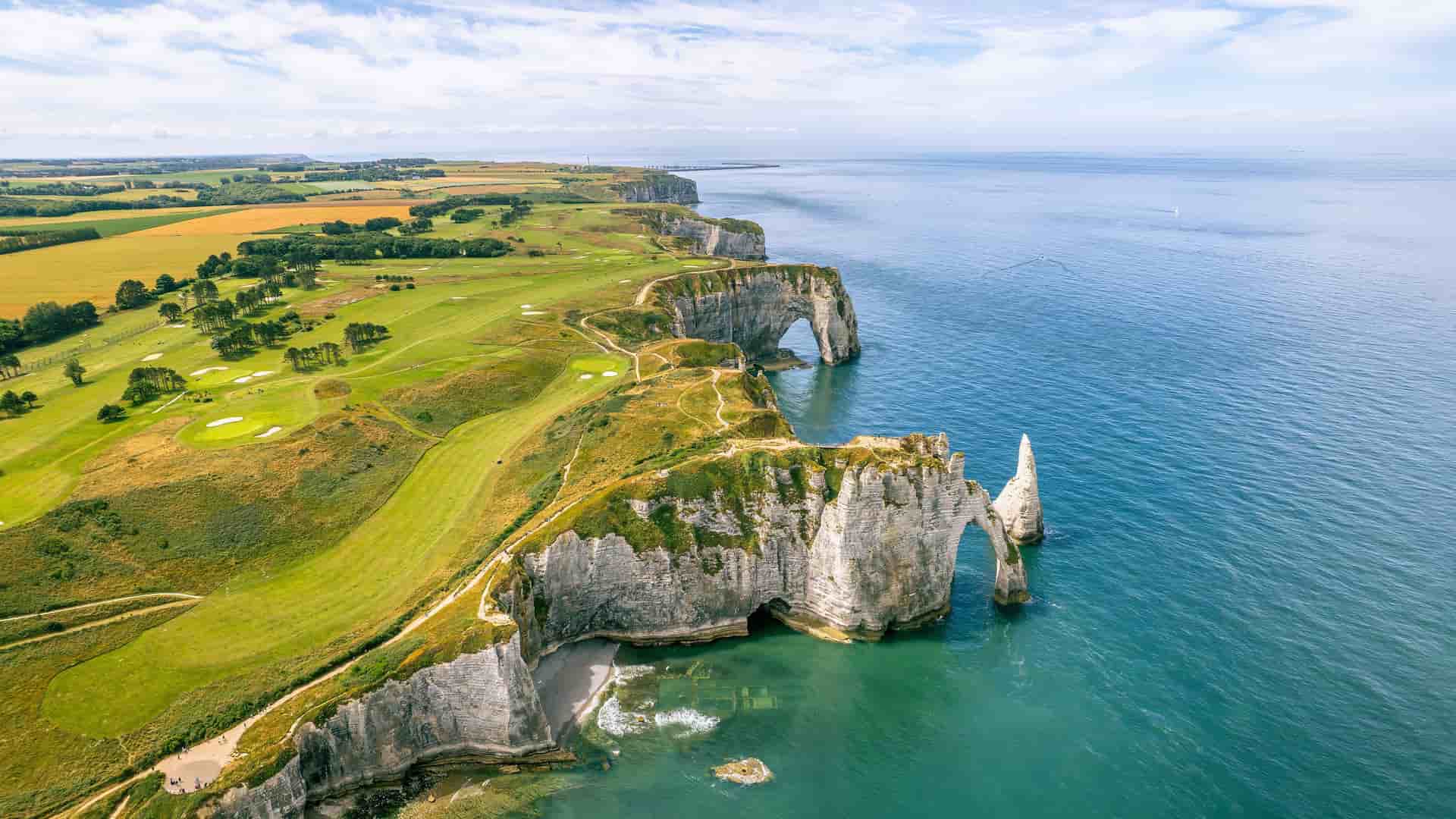 An aerial view of the iconic white chalk cliffs of Étretat, Normandy, with natural arches and a sea stack, surrounded by lush green fields and the expansive English Channel.