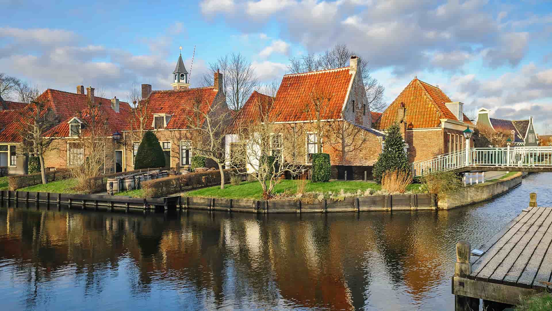 A beautiful canal in Enkhuizen, Netherlands, with historic Dutch houses with red-tiled roofs and a small footbridge reflecting perfectly in the calm water on a sunny day.